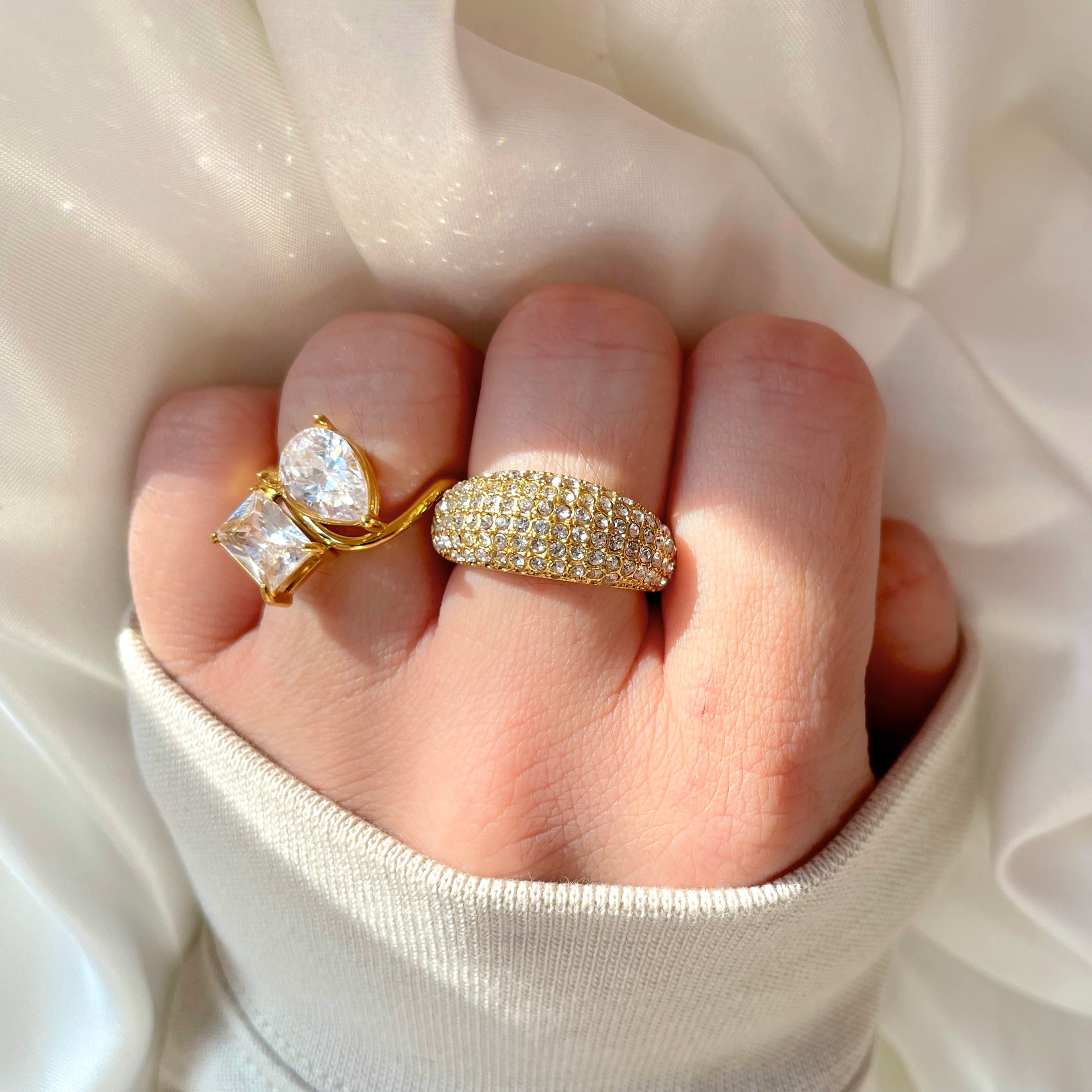 Chunky gold dome ring with clear CZ in a pave setting, stacked with a toi et moi ring on a finger against a white fabric background
