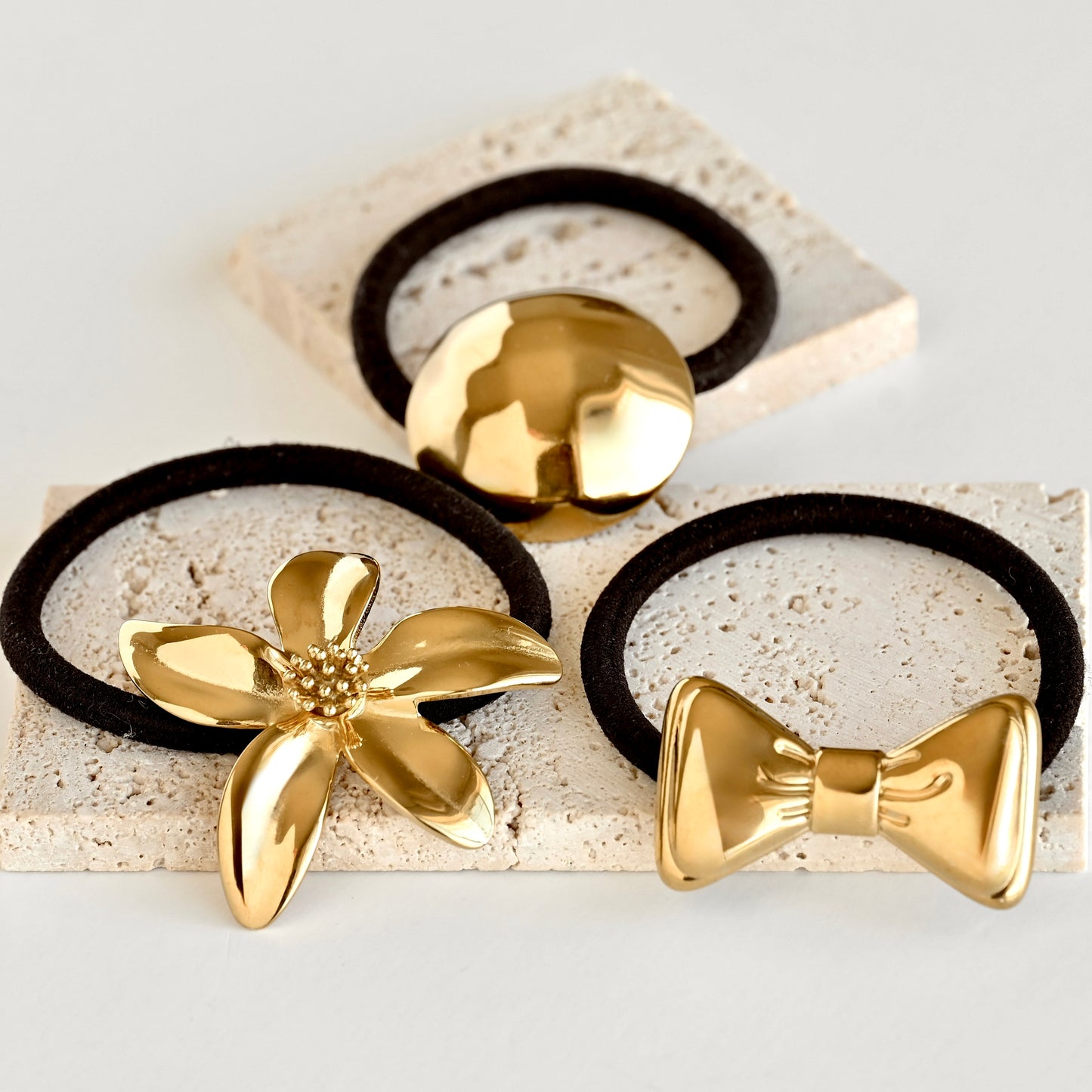 Three gold-tone hair ties with decorative metal charms are arranged on light stone blocks against a clean white background. The set includes a flower-shaped charm, a bow-shaped charm, and a round dome charm, all attached to black elastic bands.