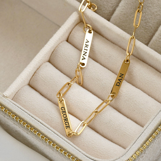 A gold paperclip chain bracelet with three engraved name tags (ARINA, GEORGE, and DAN) displayed inside a beige jewelry box.