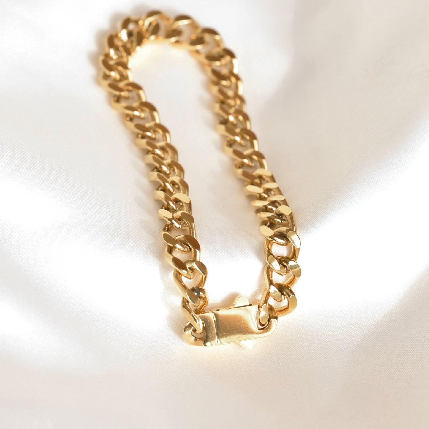 A gold-toned Cuban link chain bracelet with a lobster clasp, displayed on a white satin fabric background.