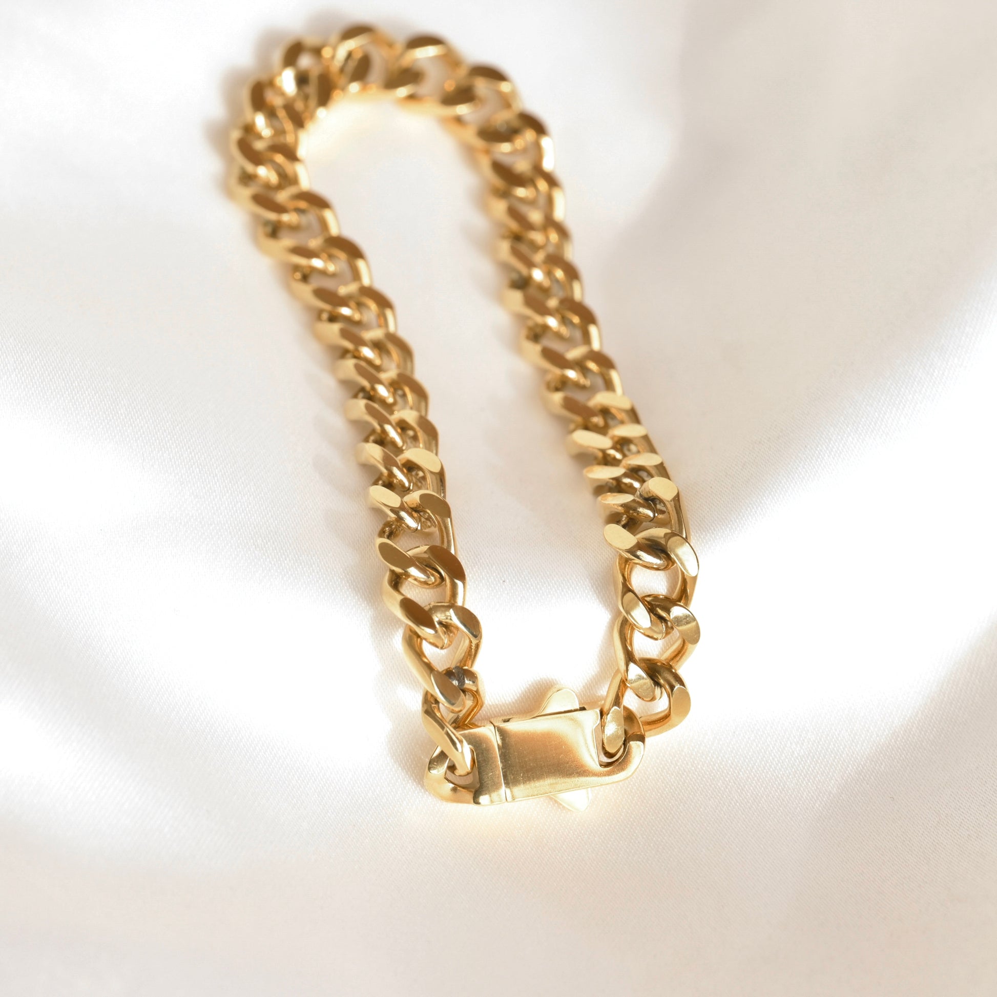 A gold-toned Cuban link chain bracelet with a lobster clasp, displayed on a white satin fabric background.