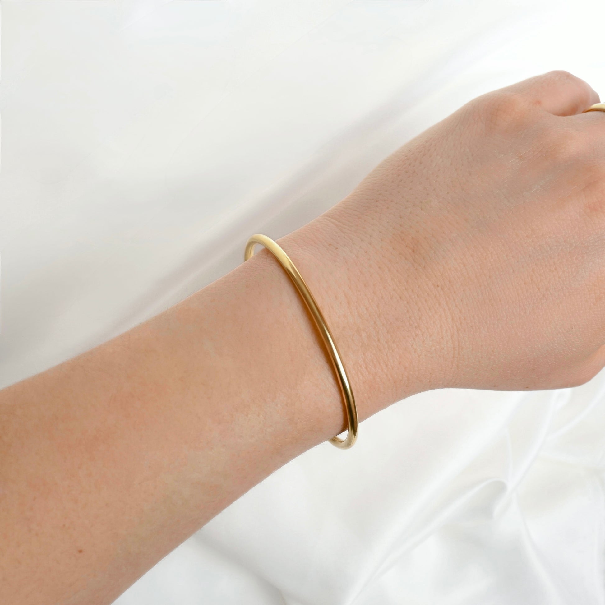 A minimalist, thin gold bangle bracelet worn on a person's wrist, shown against a soft white fabric background.