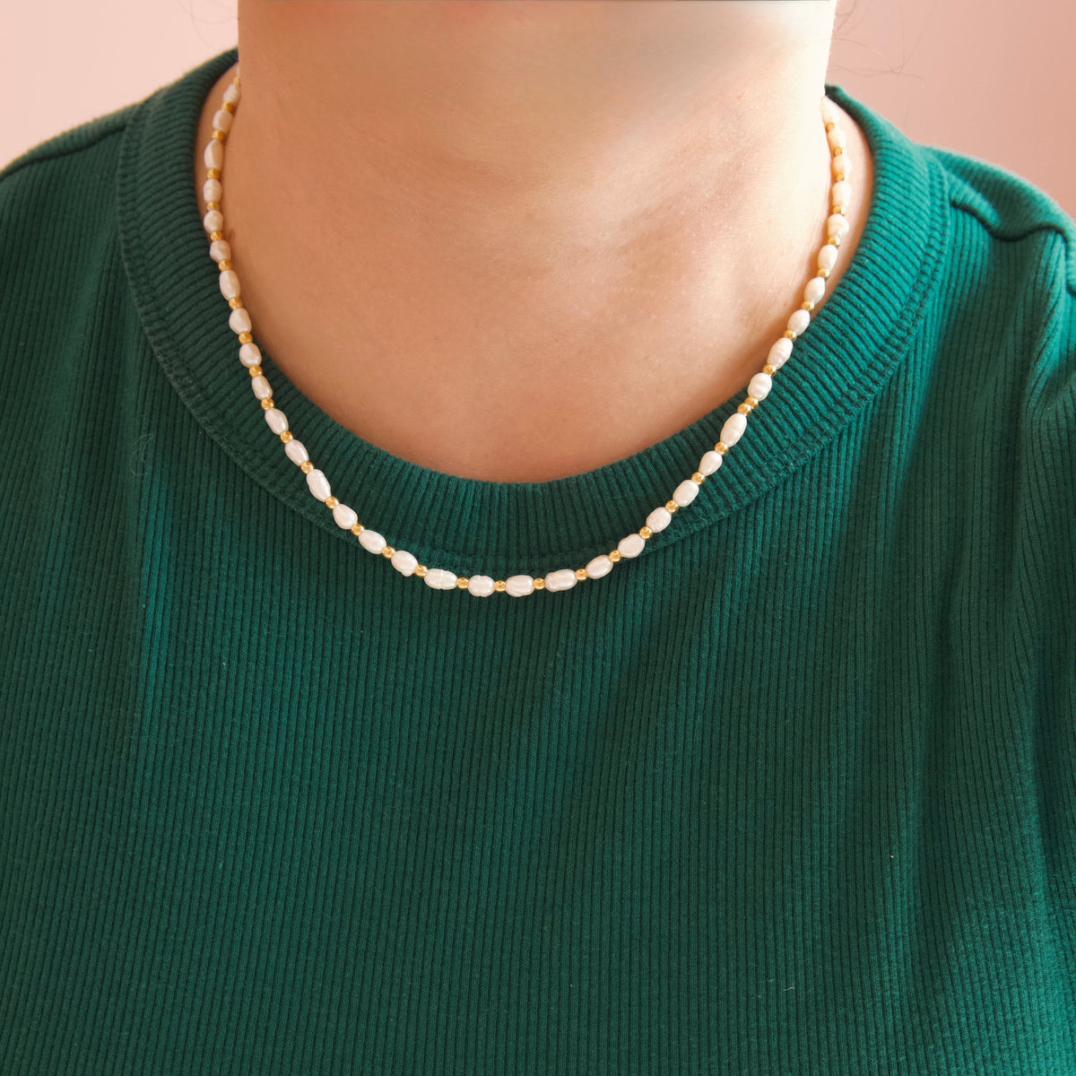 A close-up of a person wearing a short strand of cream-colored pearl bead necklace over a dark green ribbed knit top.