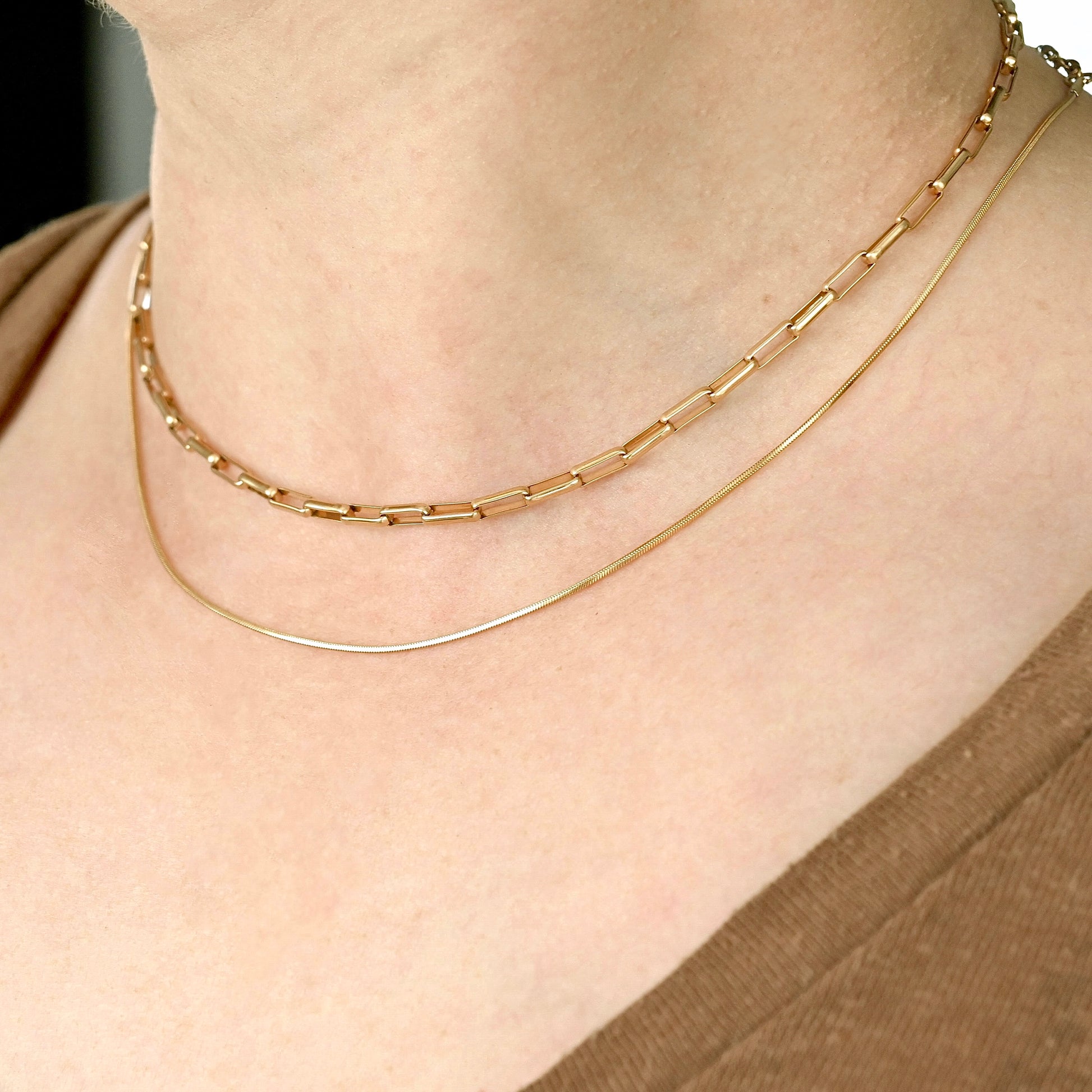 Two gold necklaces, one longer snake and one shorter bold paperclip,  stacked on a person's neck 