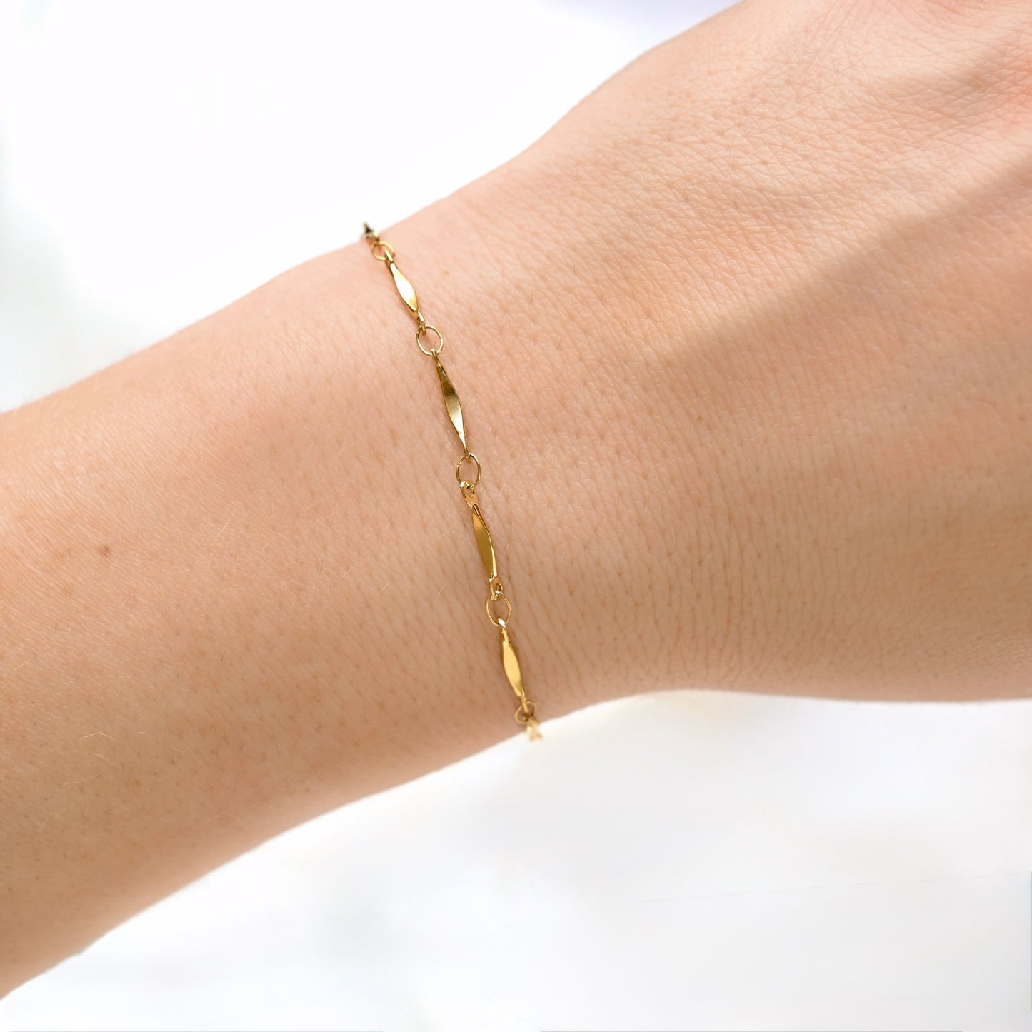 A delicate gold chain bracelet with elongated oval links, shown worn on a person's wrist against a bright white background.