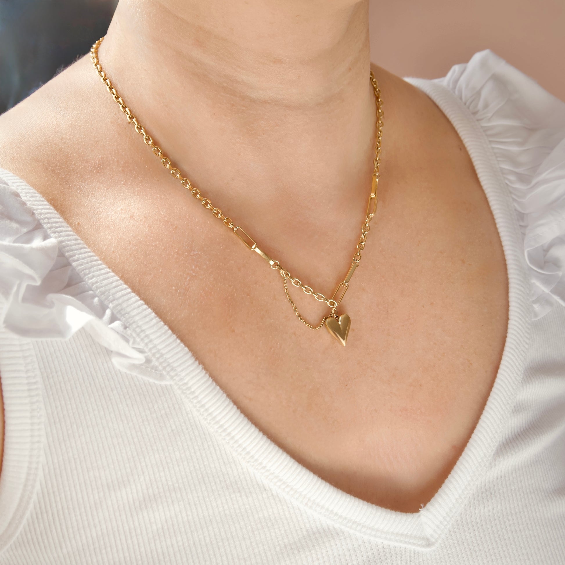 A close-up of a model wearing a delicate gold chain necklace with a small polished heart pendant, styled with a white ruffled top.