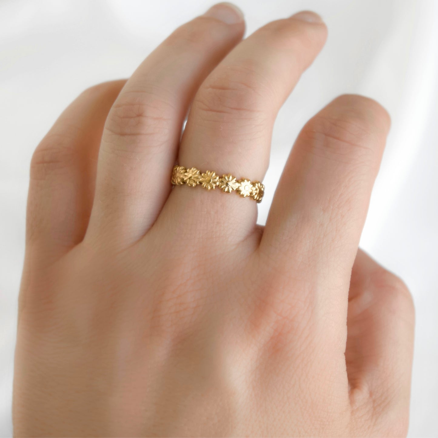 Dainty gold daisy chain band on a woman's middle finger against white background.