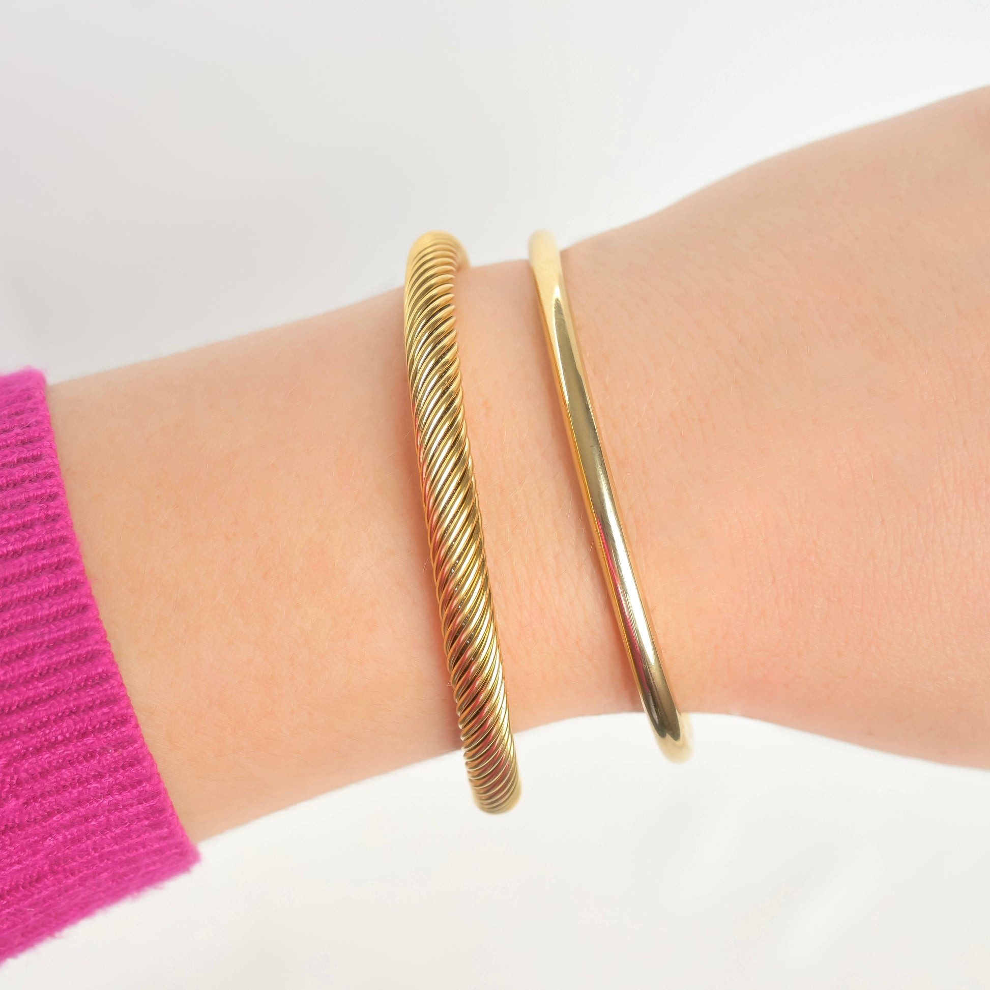 Gold twisted cable and high-polish cuff bracelets on a wrist with a pink sleeve against a white background