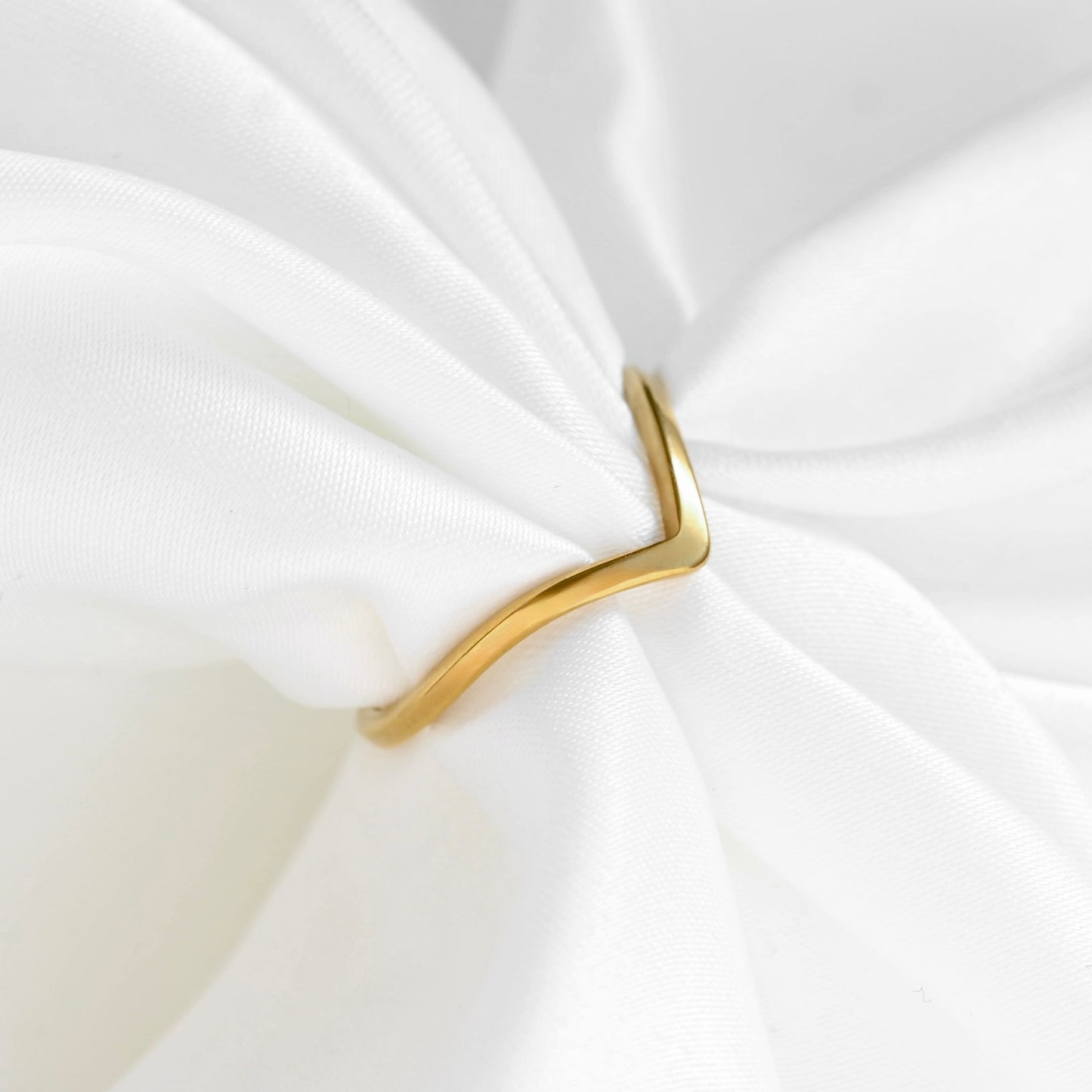 A slim, polished gold ring with a unique wavy, open V-shaped design, shown tied around a soft white fabric napkin.