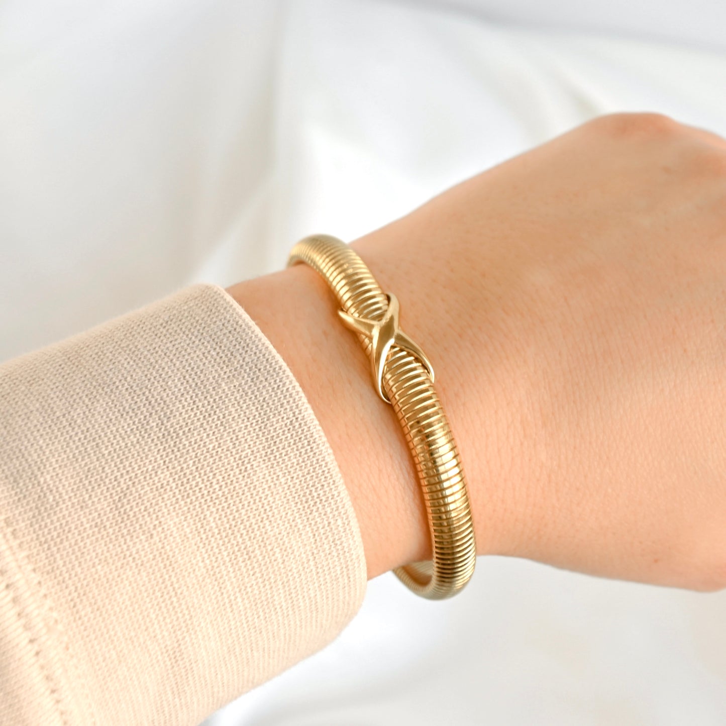 Dainty gold tubogas bangle bracelet with a love knot on a wrist with a plain background