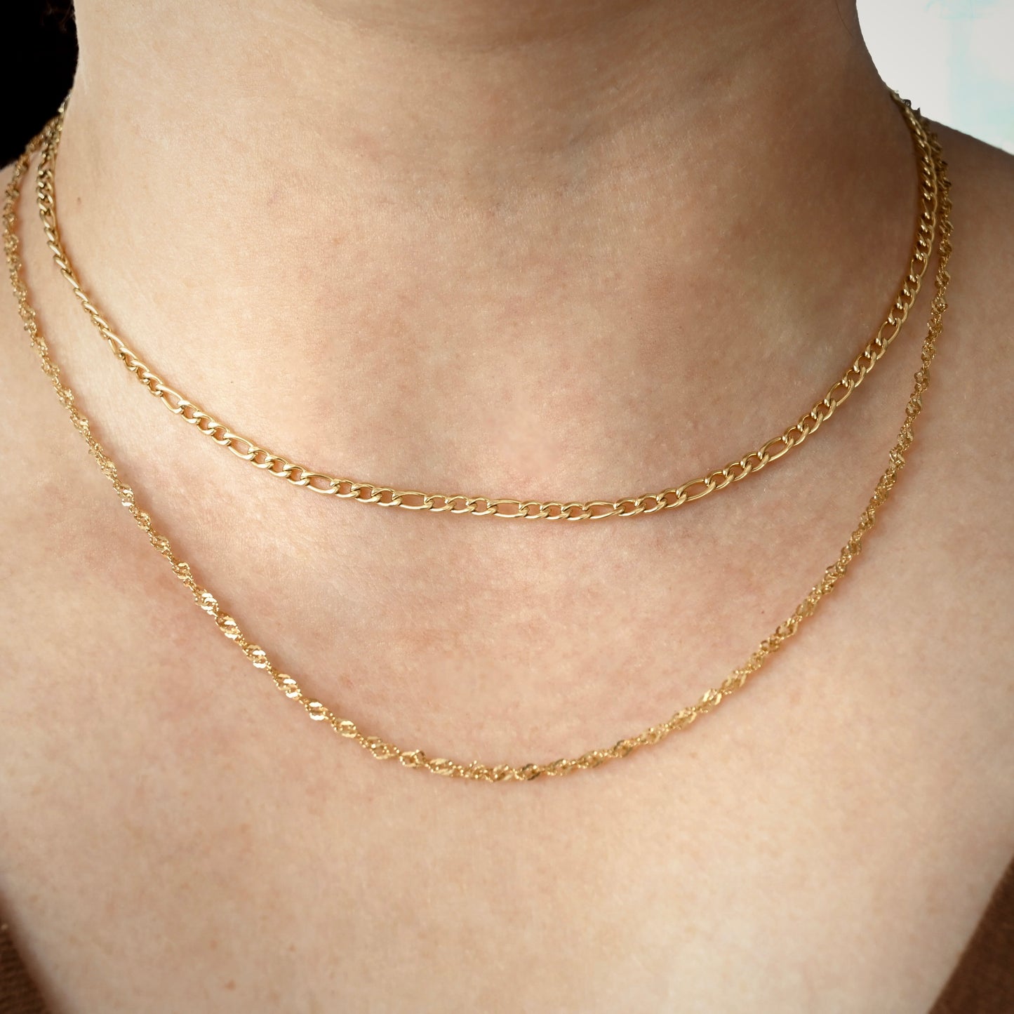 Gold figaro chain layered with a  longer singapore chain on a person's neck with a neutral background