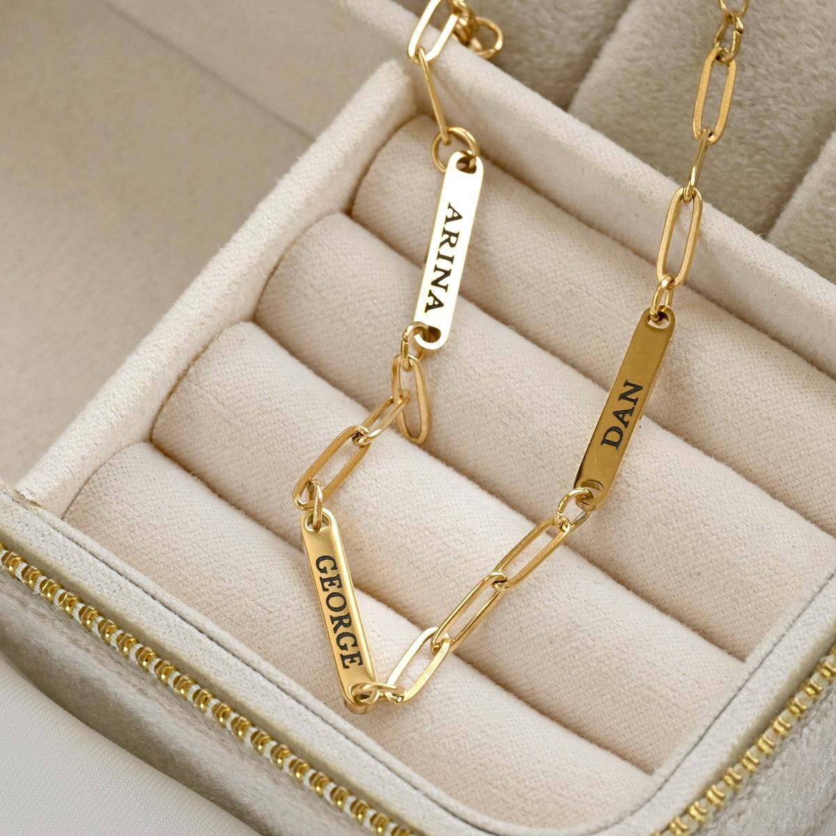 A gold chain necklace with three engraved name tags (ARINA, GEORGE, and DAN) displayed inside a beige jewelry box.