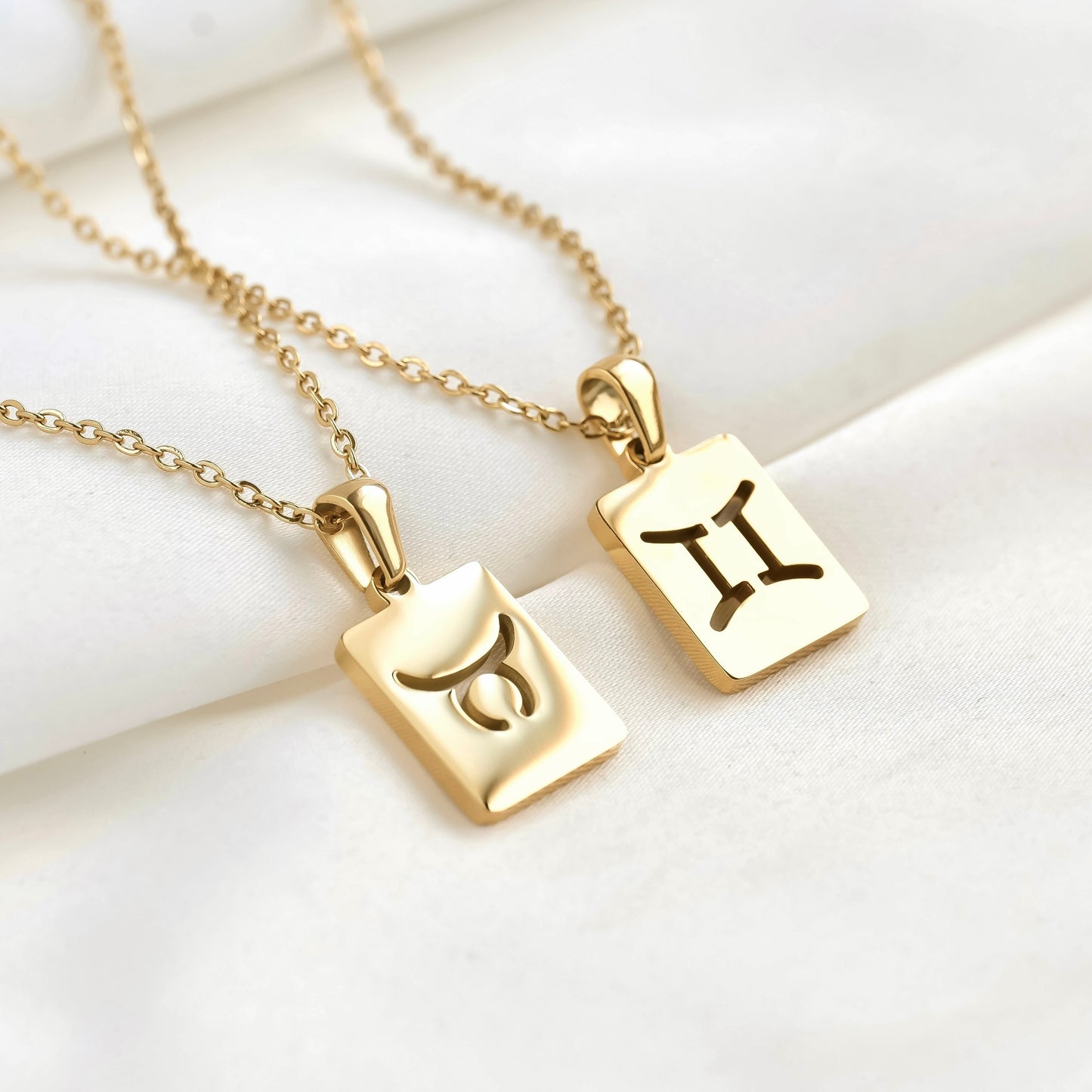 Two gold necklaces with rectangular pendants featuring astrological symbols on a white surface.