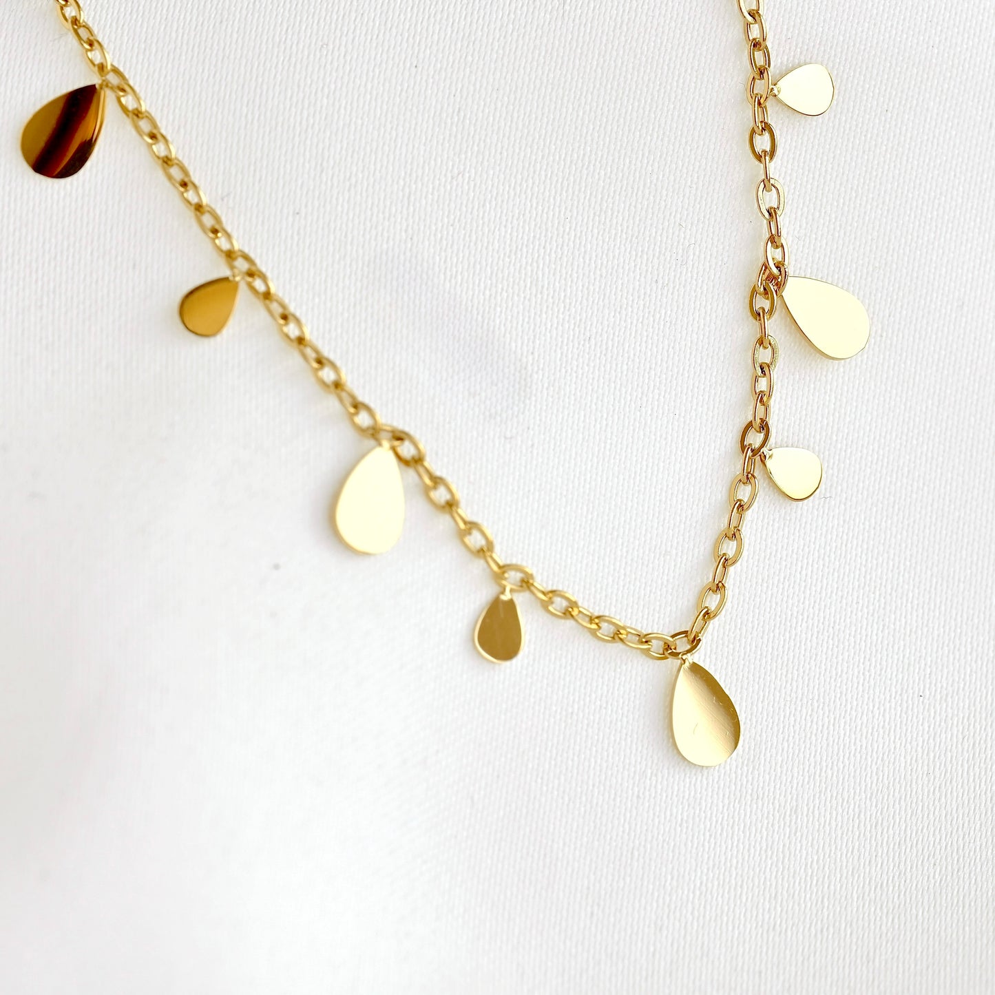 A gold chain necklace with multiple flat, teardrop-shaped gold charms of varying sizes, displayed on a textured white fabric background.