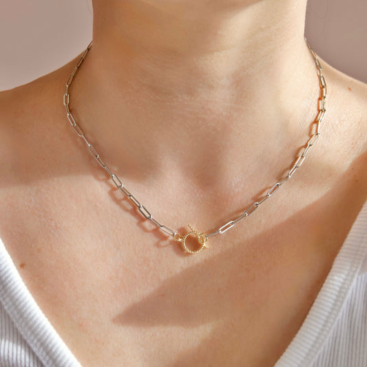 A close-up of a person's neck and upper chest wearing a delicate paperclip chain necklace with a small gold ring clasp.