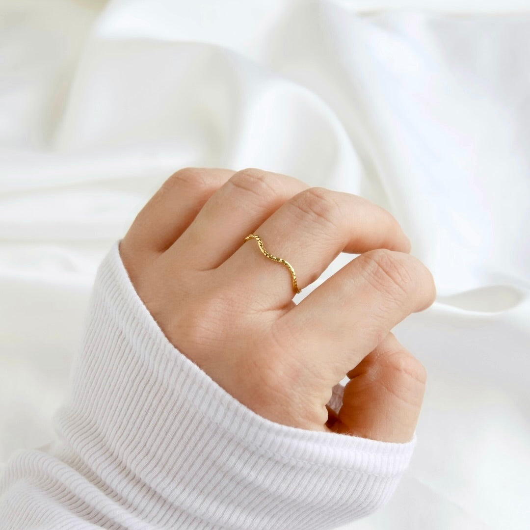 A close-up of a hand wearing a delicate, wavy gold ring, dressed in a white ribbed knit sweater against a soft, draped white fabric background.
