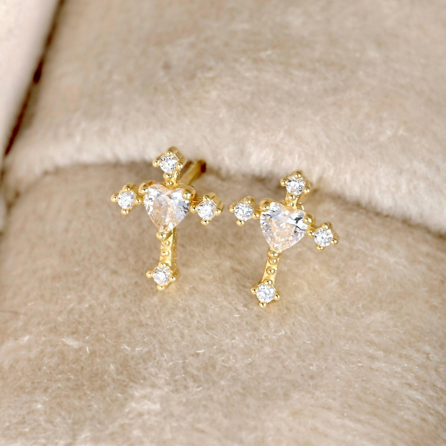 A pair of gold cross stud earrings with heart-shaped clear crystals and small round crystal accents, displayed on a soft beige textured surface.