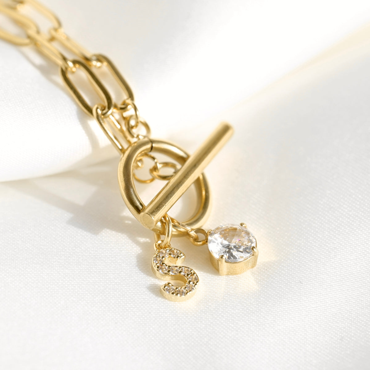 Gold-tone toggle clasp necklace with a paperclip chain, a dangling letter S charm, and a clear round crystal charm, displayed on a white fabric background.