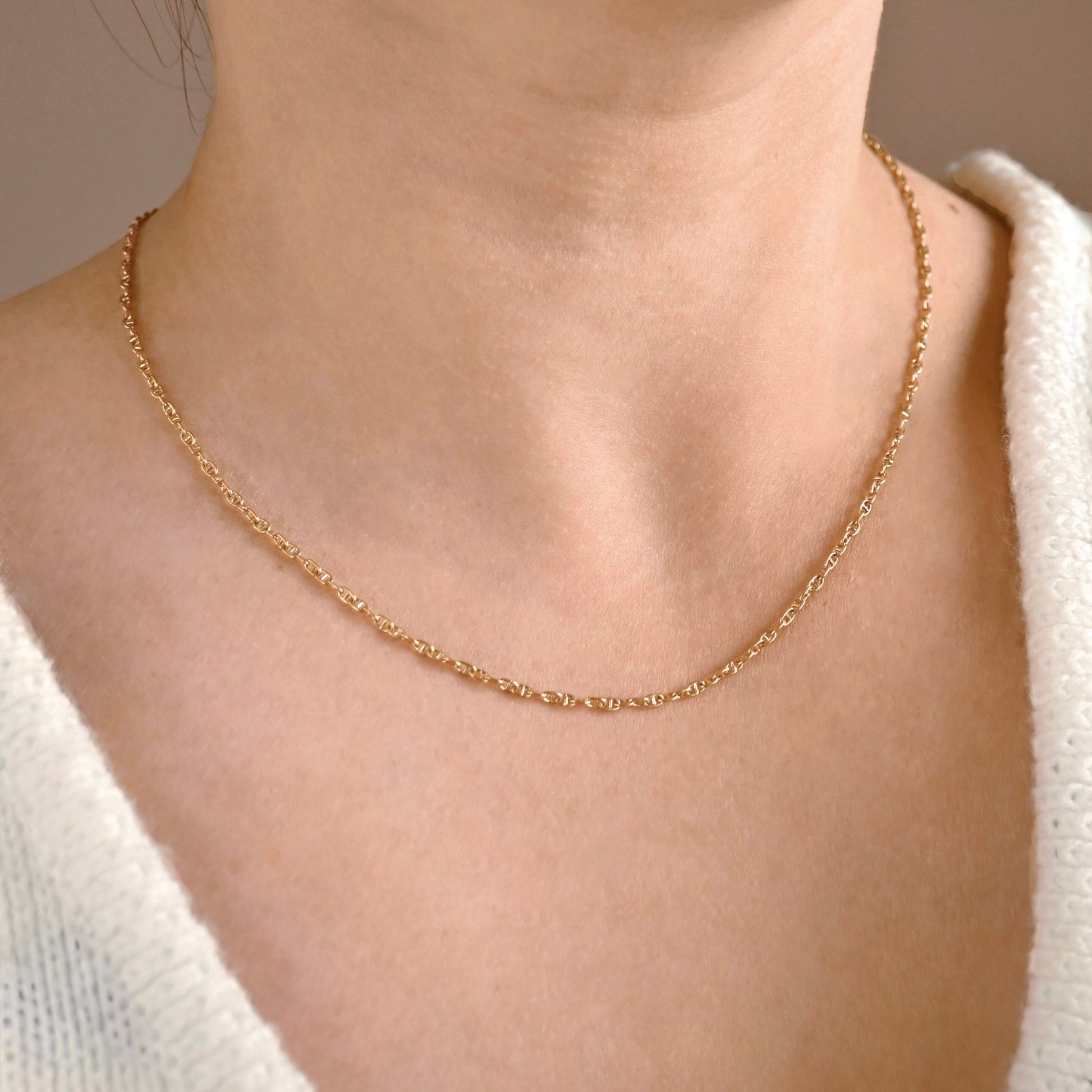 A close-up of a person's neck and upper chest wearing a delicate gold chain necklace with a small mariner chain link design, paired with a white knit top.