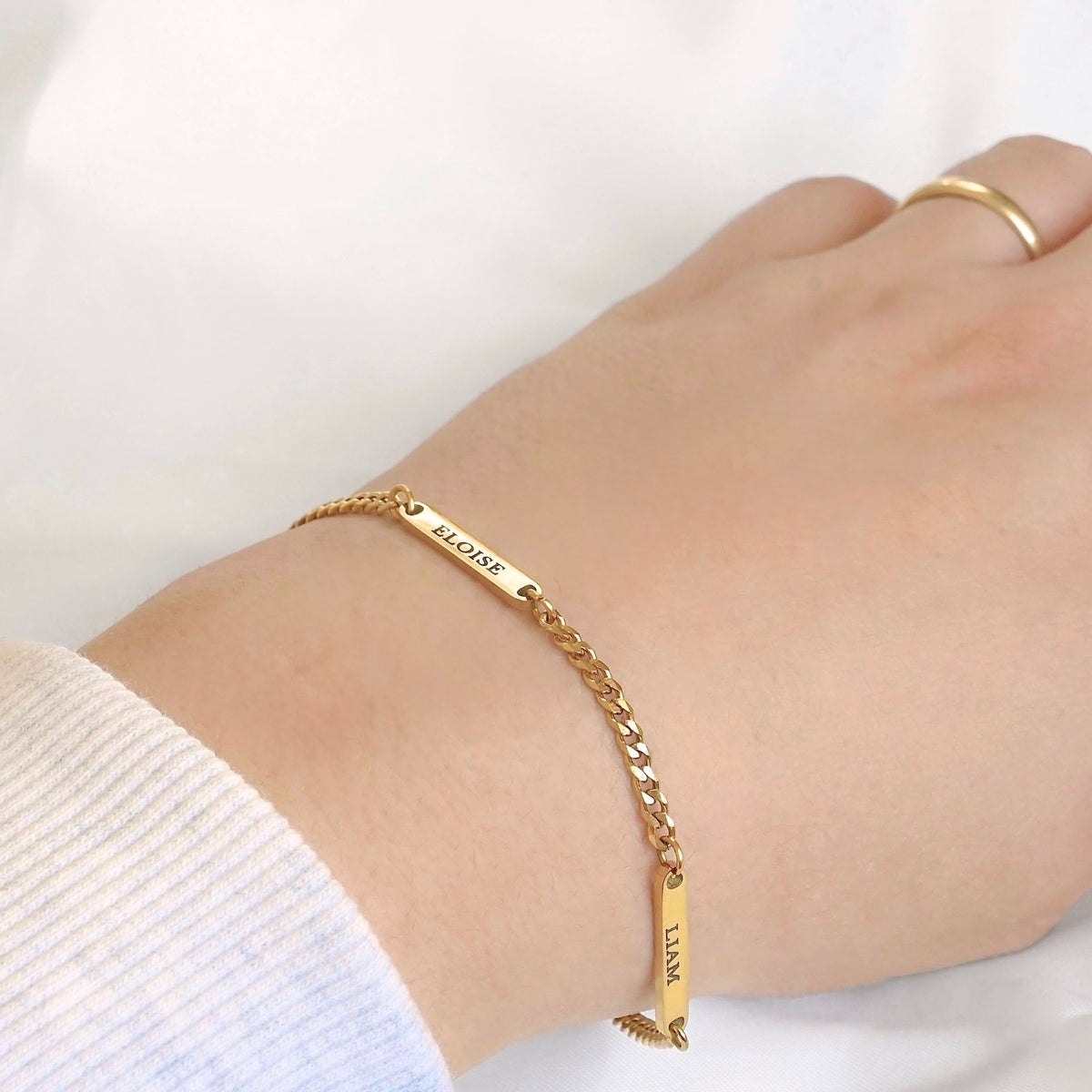 A delicate gold CURB chain bracelet with engraved nameplates worn on a person's wrist, shown against a soft white background with a white ribbed sleeve visible.
