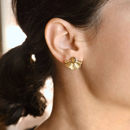 A close-up of a person's ear wearing a small gold fan-shaped stud earring with a round amber-brown center stone.