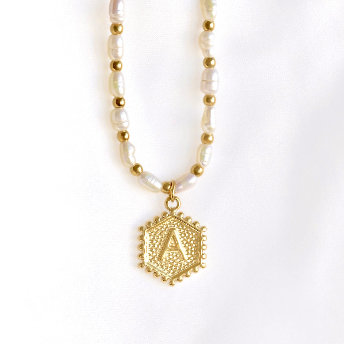 A gold and white beaded necklace featuring a textured gold hexagonal pendant with an initial 'A' monogram, displayed on a clean white background.