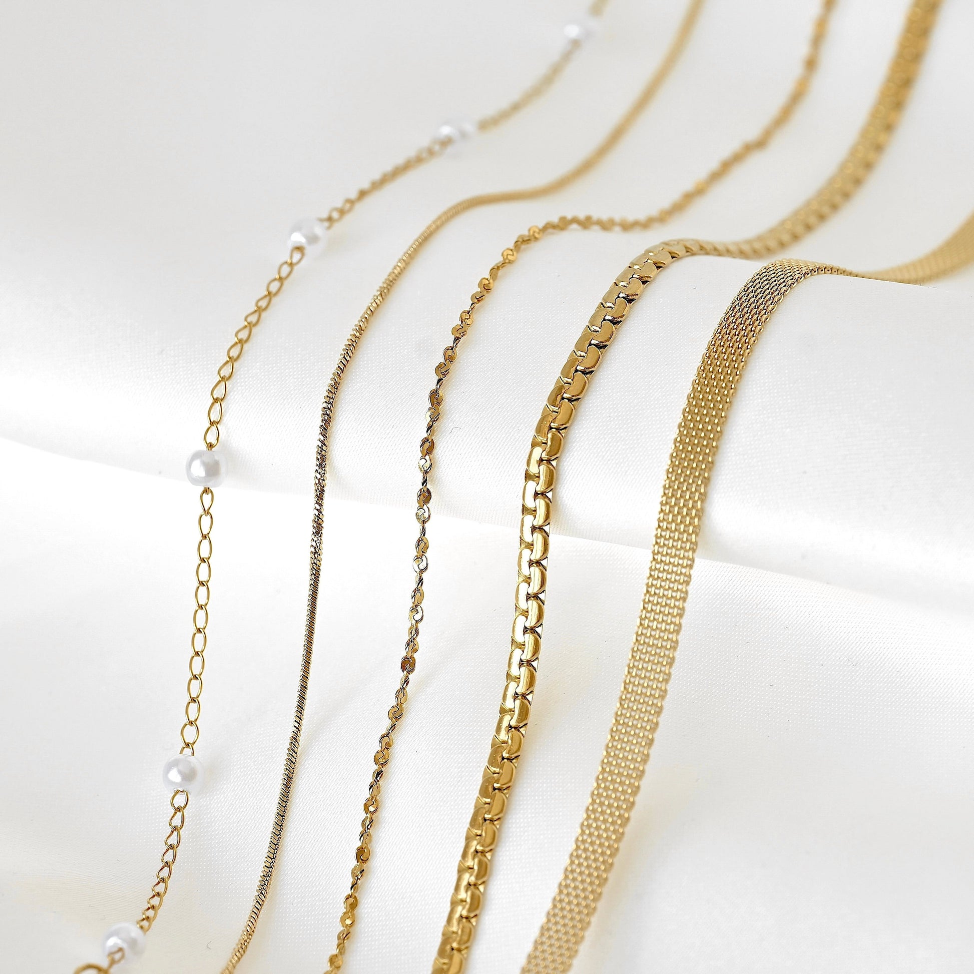 Five gold-tone chain necklaces with different link styles and pearl accents, arranged diagonally on a white satin fabric background.