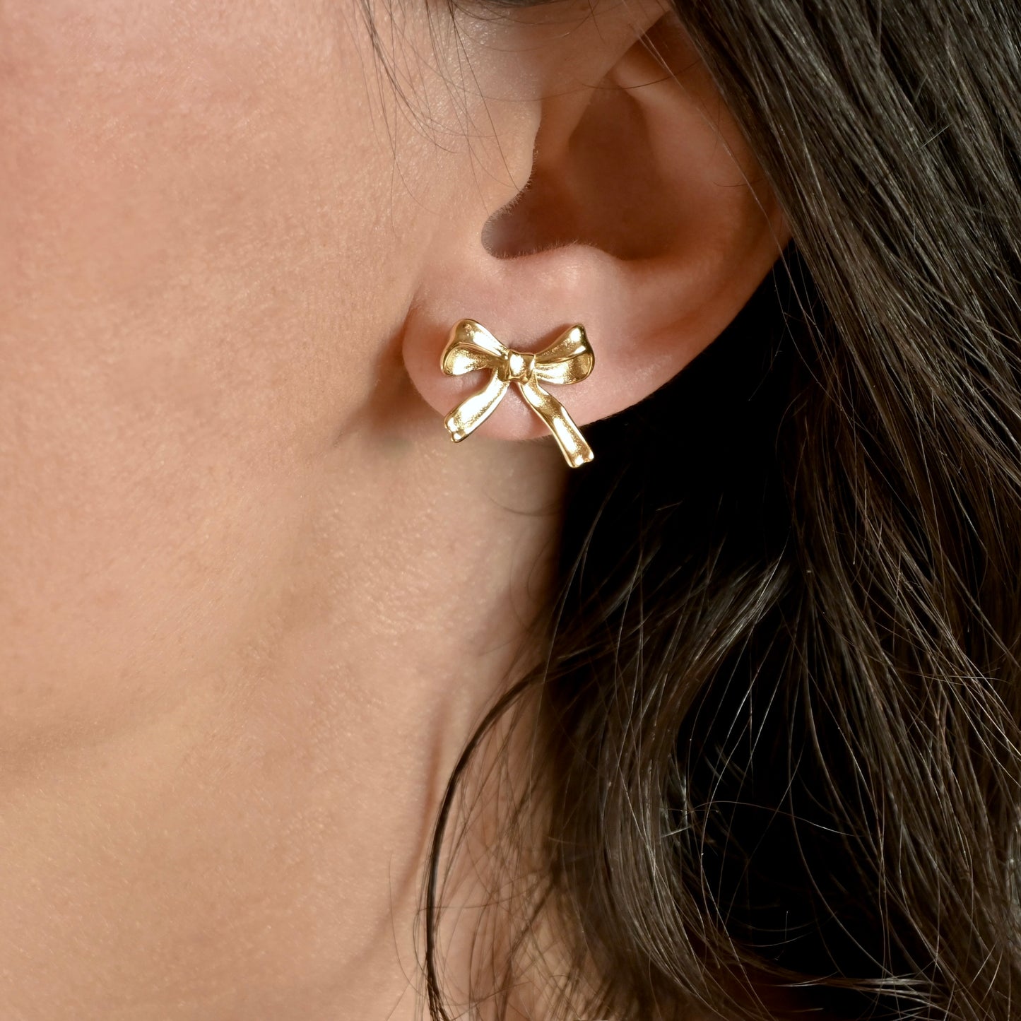A close-up of a person's ear wearing a small polished gold bow stud earring.