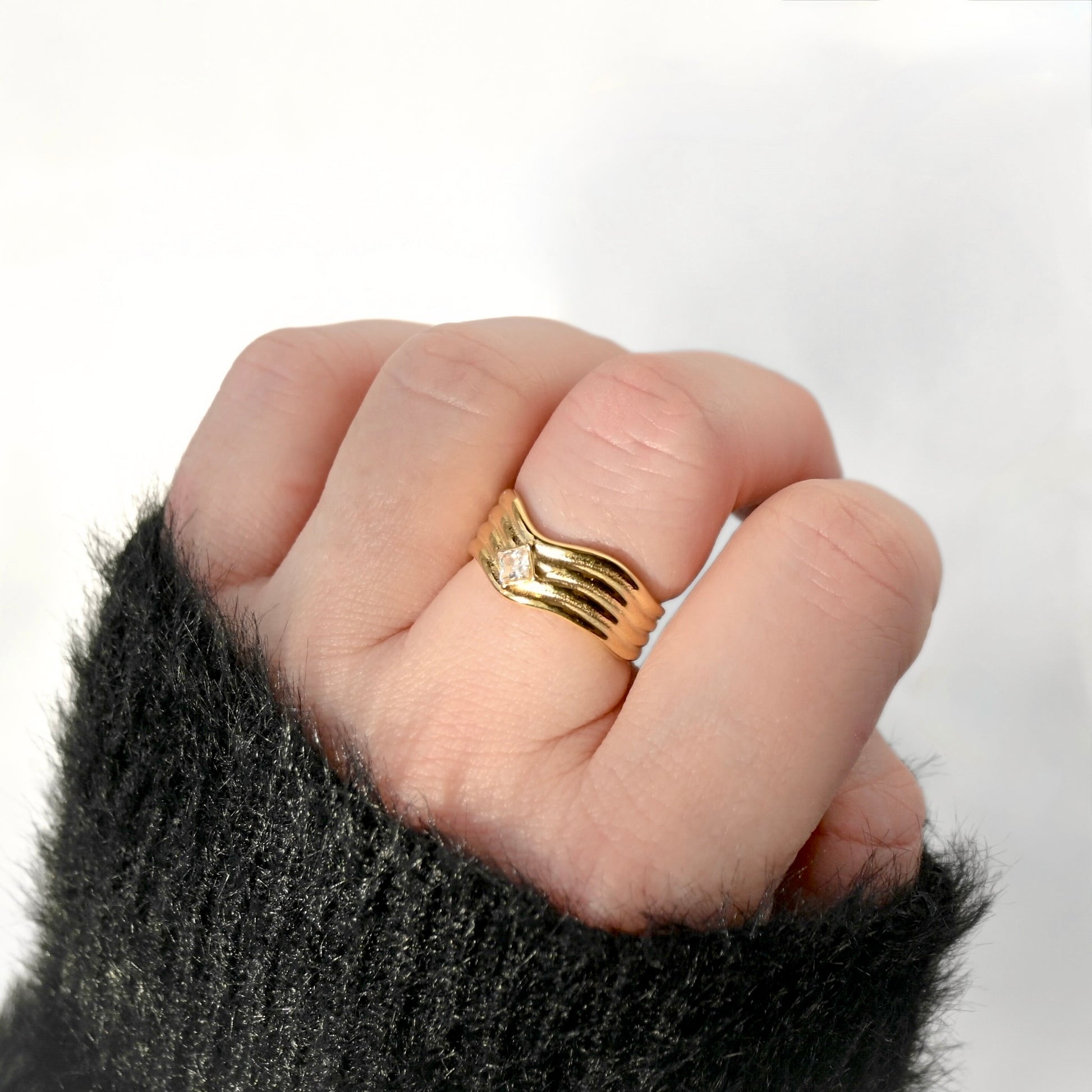 A gold ring with a wavy, layered design with a central clear diamond shaped stone,  worn on a person's finger, shown against a white background with a black fuzzy sleeve visible.