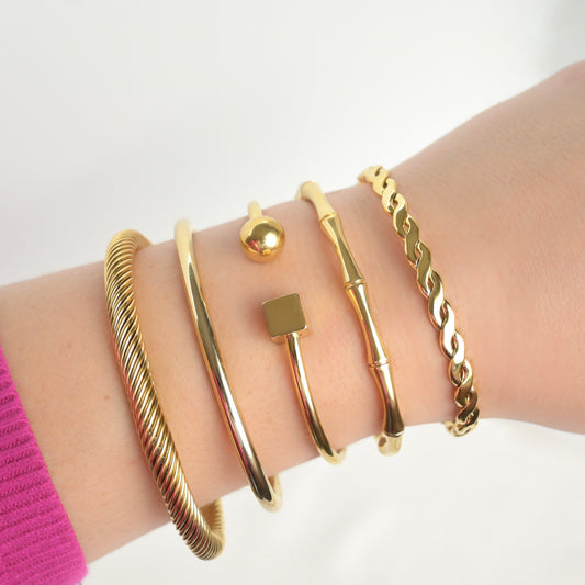 Stack of gold cuff bracelets inlcuding a twisted rope, tube, cube and ball, bamboo and braided designs, on a wrist against a white background