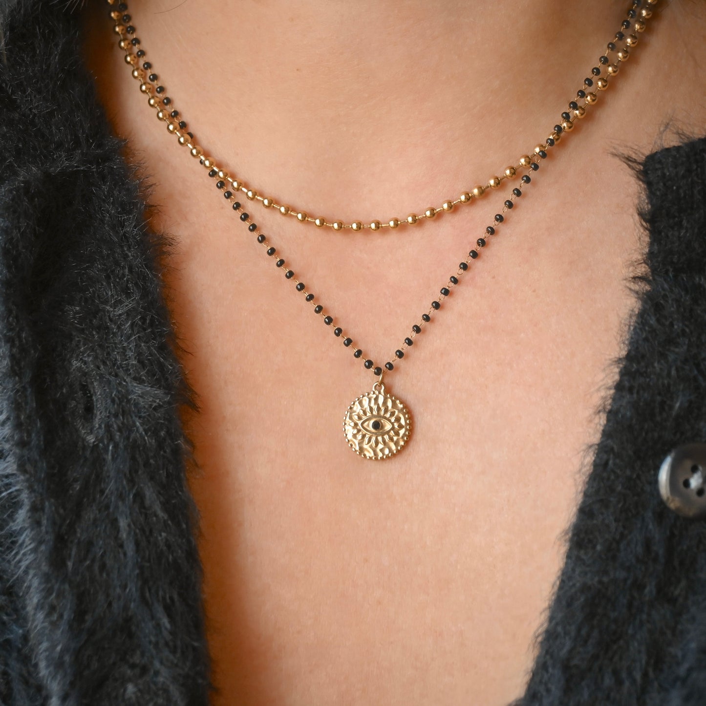A close-up of a person wearing a layered necklace set. The set features one delicate gold chain with a textured circular sunburst pendant and a second black beaded chain layered over it.
