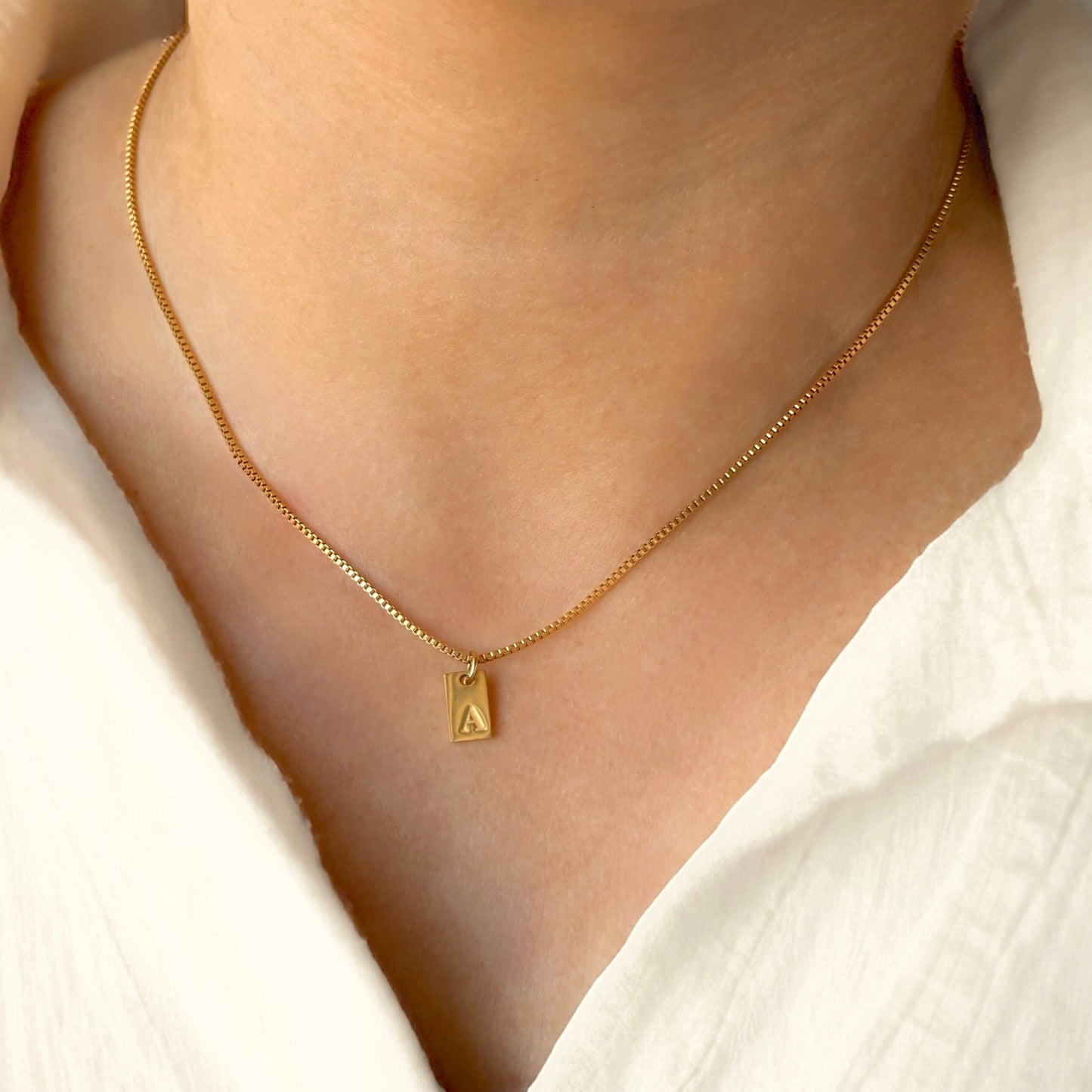 Gold box chain necklace with a tag initial pendant featuring the letter 'A' on a person wearing a white blouse.