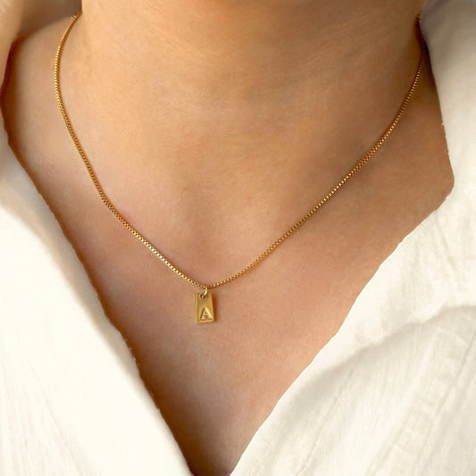 Gold box chain necklace with a tag initial pendant featuring the letter 'A' on a person wearing a white blouse.