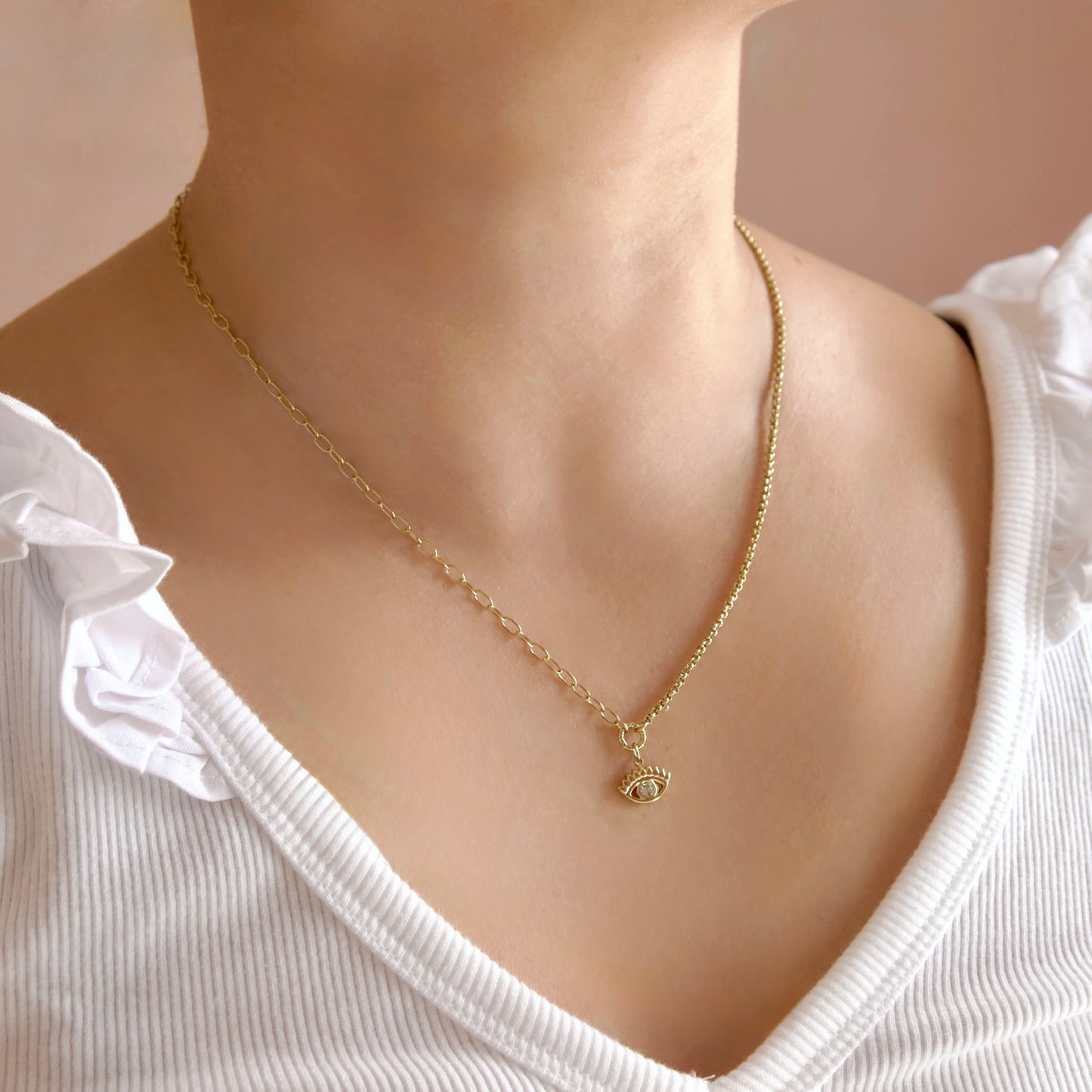 A close-up of a person wearing a delicate gold chain necklace with a small evil eye pendant, paired with a white ribbed top.