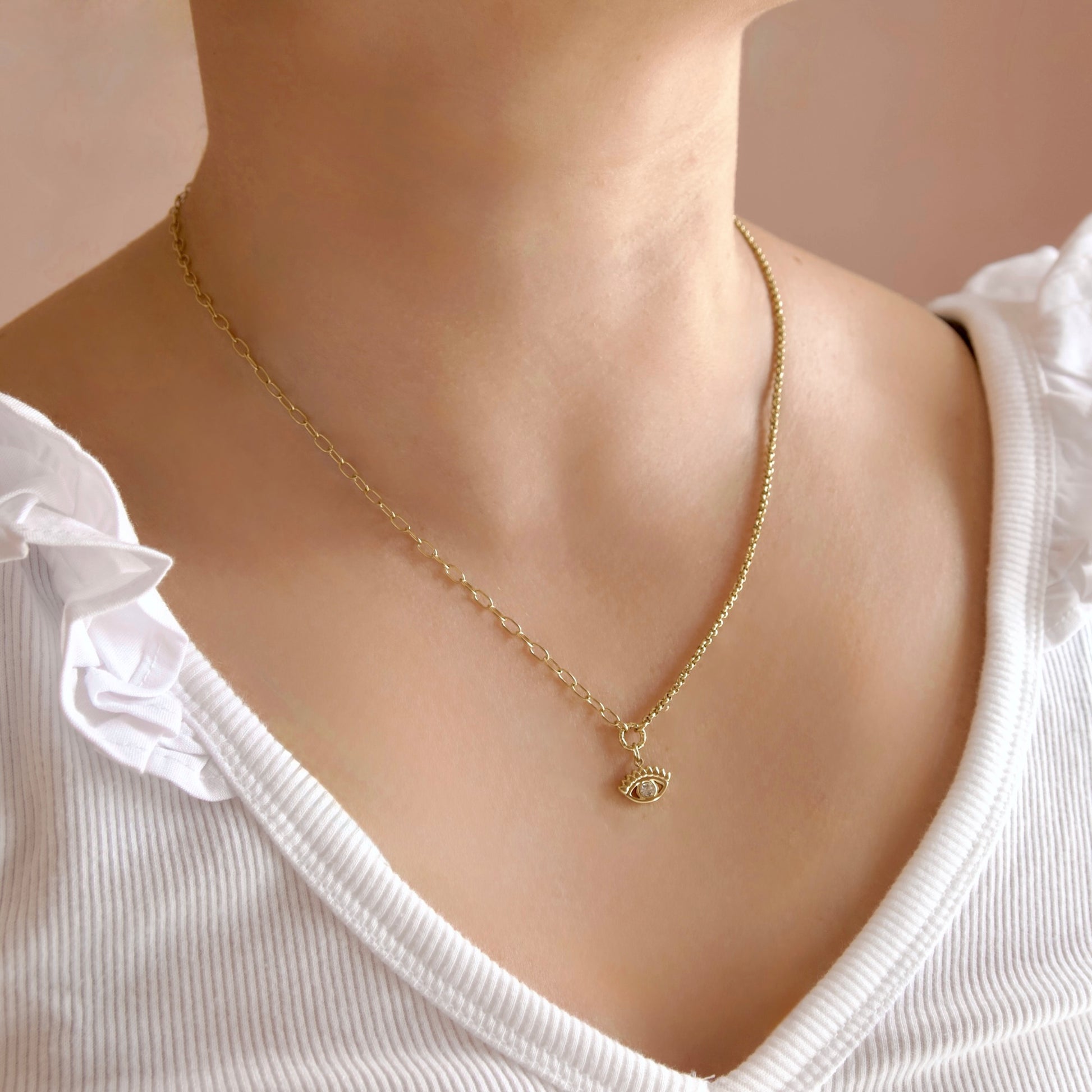 A close-up of a person wearing a delicate gold chain necklace with a small evil eye pendant, paired with a white ribbed top.