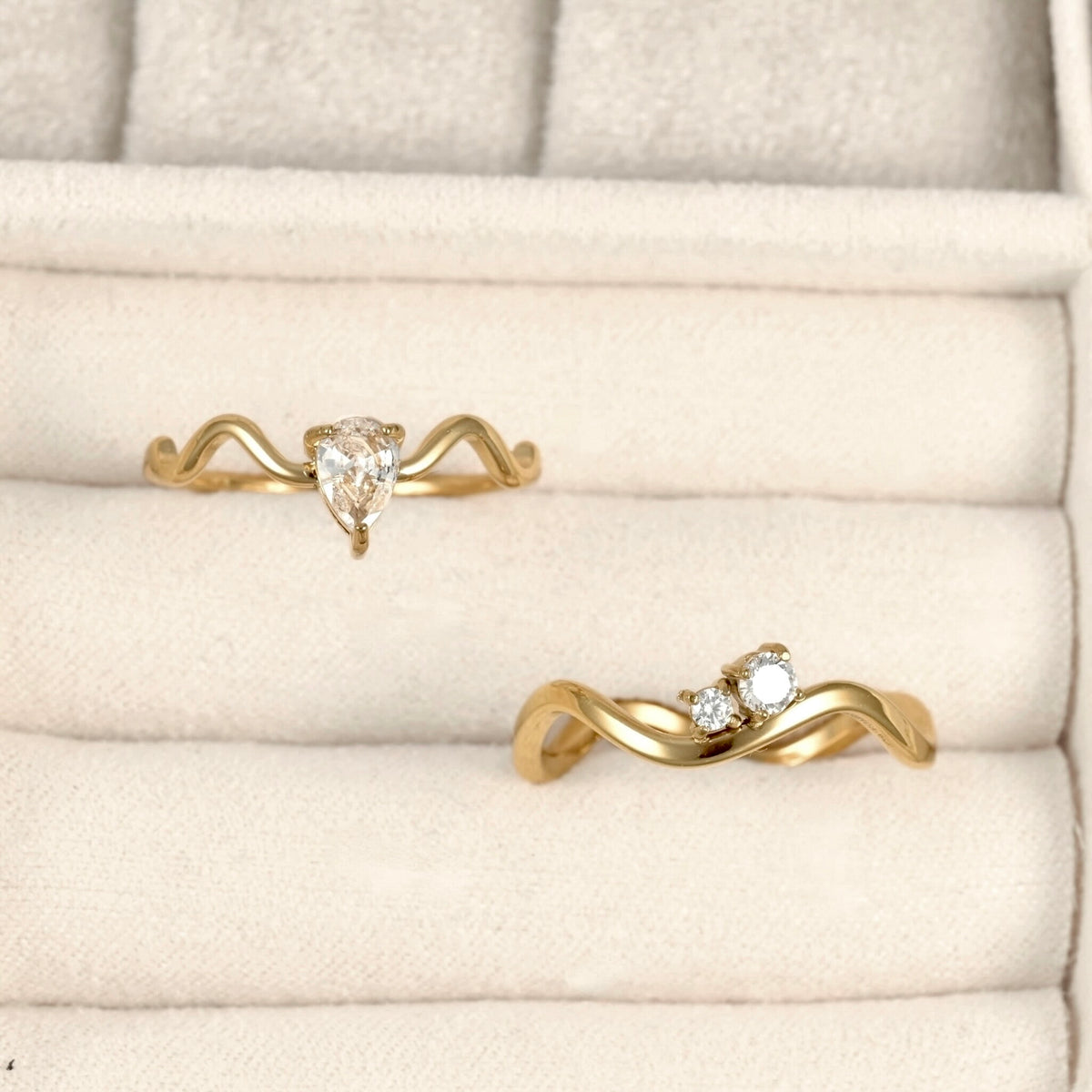 Two gold rings displayed on a cream-colored ring box. One ring features a pear-cut clear gemstone, and the other features a small cluster of clear gemstones on a wavy band.