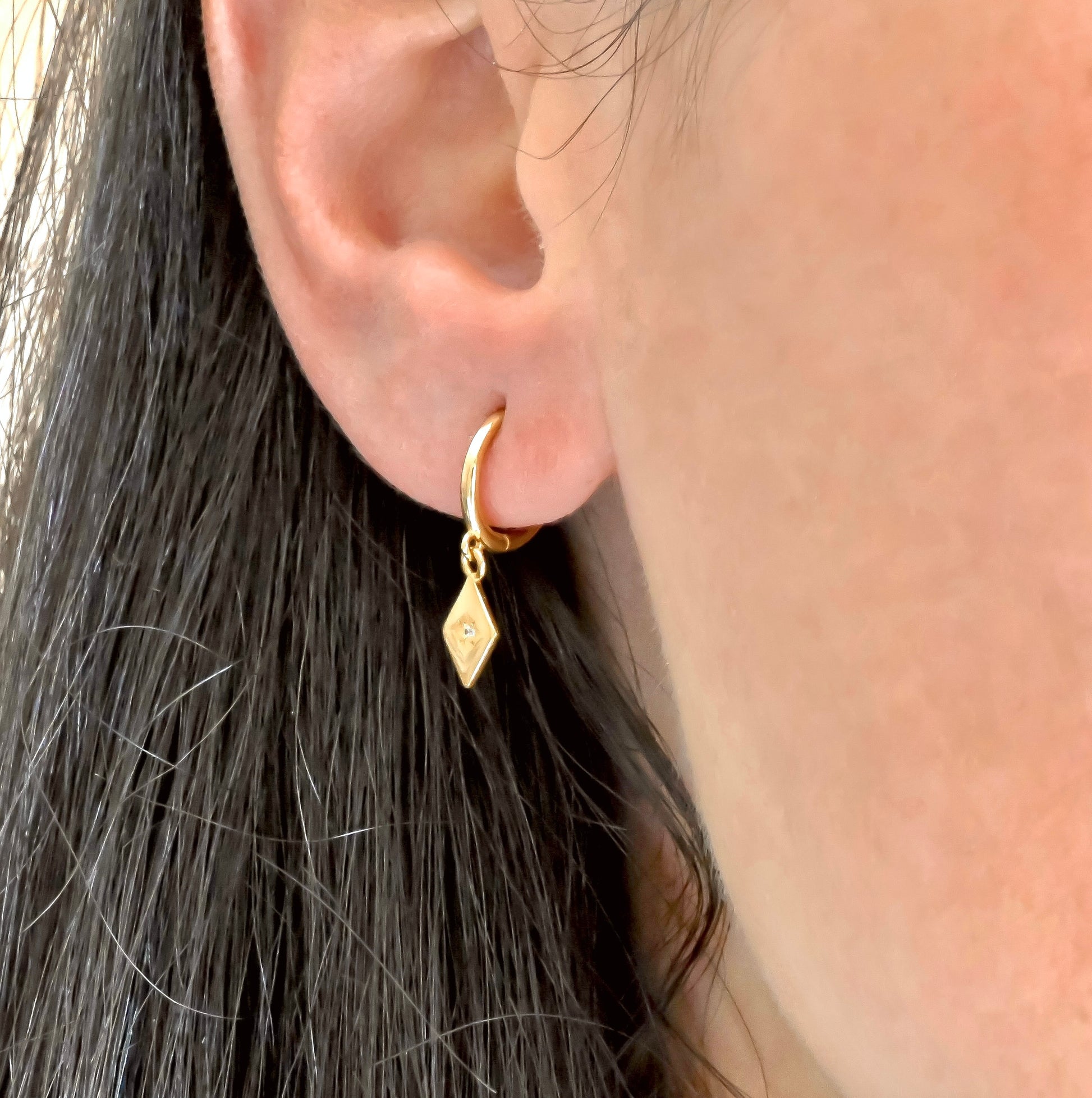Gold rhombus charm hoop earring worn by a person with dark hair.