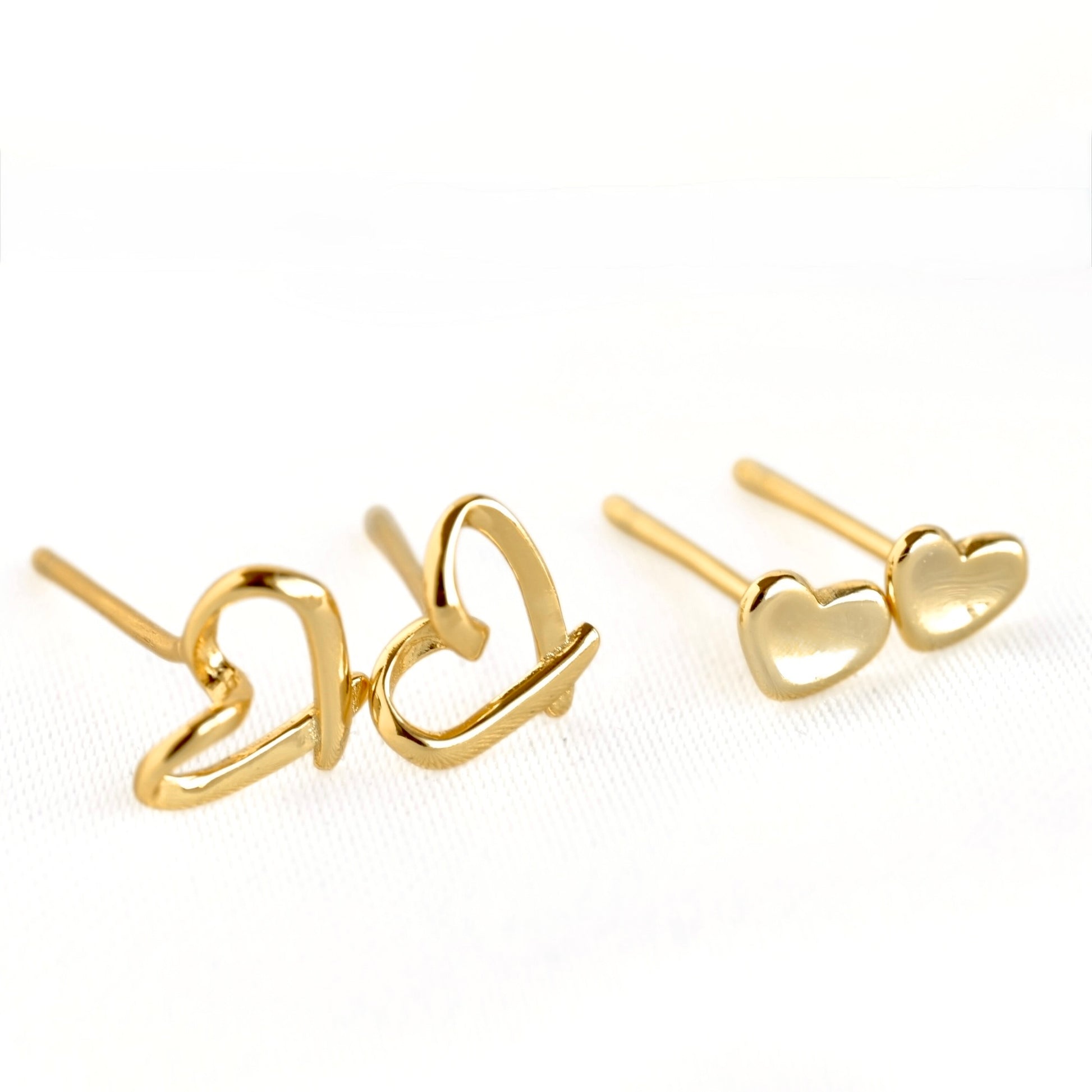 A pair of gold heart stud earrings, with one earring showing an open heart outline design and the other showing two small solid heart charms, photographed on a clean white background.