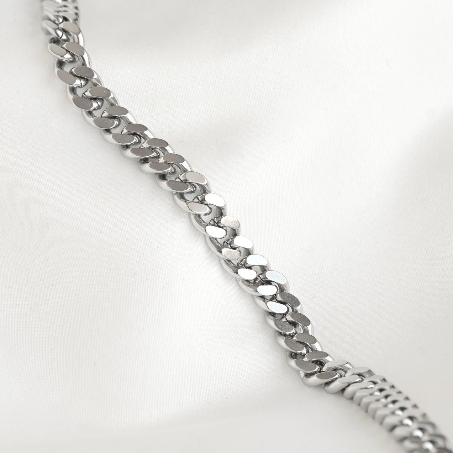 A silver-toned metal curb link chain bracelet displayed diagonally on a soft white fabric background.