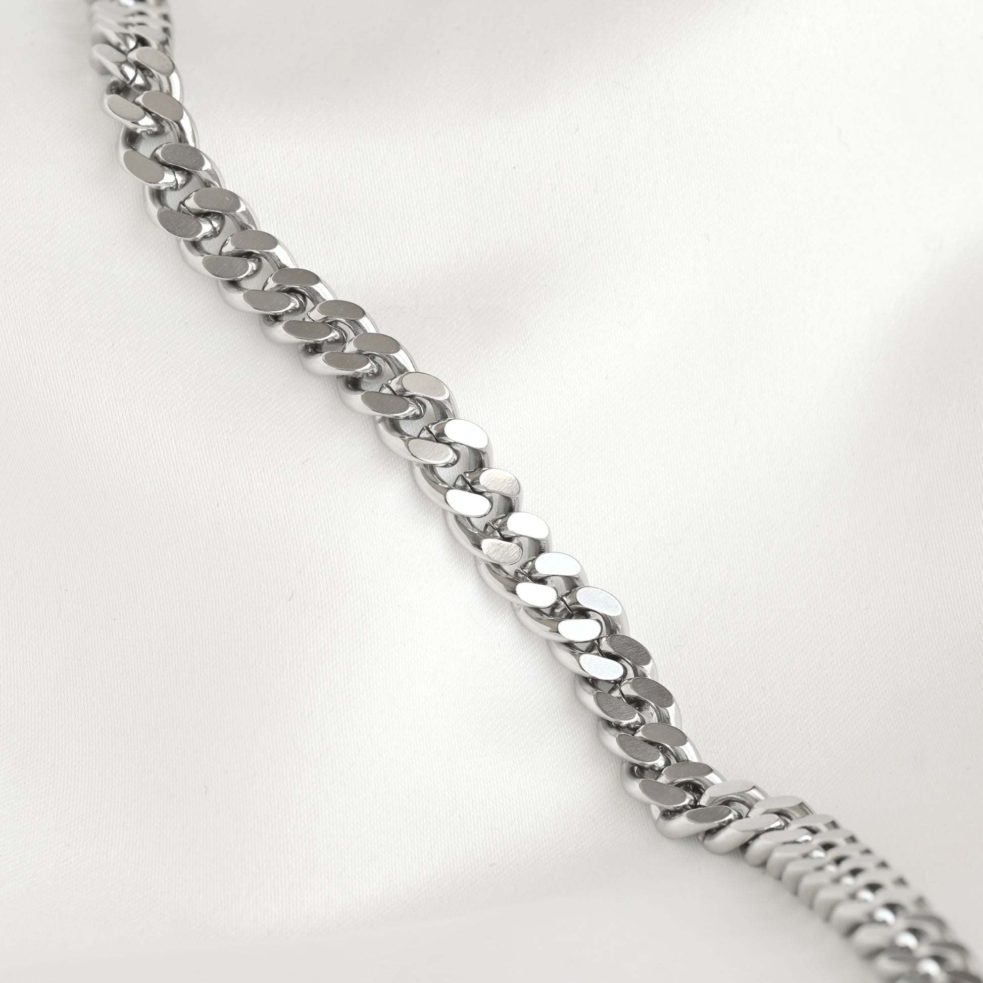 A silver-toned metal curb link chain bracelet displayed diagonally on a soft white fabric background.