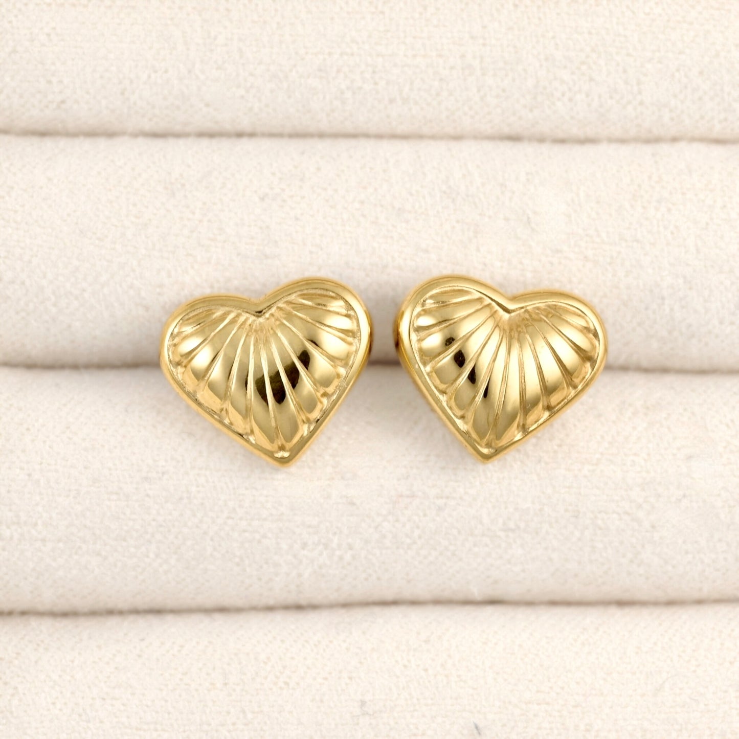 A pair of gold heart-shaped stud earrings with a textured, ribbed surface, displayed on a cream-colored textured fabric background.