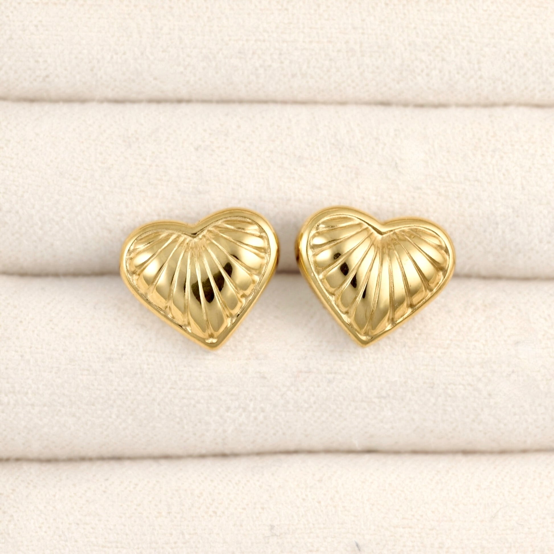 A pair of gold heart-shaped stud earrings with a textured, ribbed surface, displayed on a cream-colored textured fabric background.