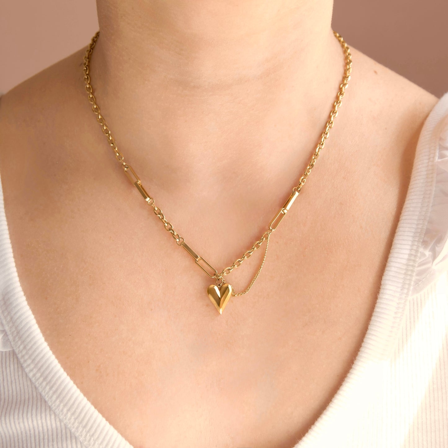 A close-up of a person wearing a delicate gold chain necklace with a small polished heart pendant, paired with a white ribbed top against a soft beige background.