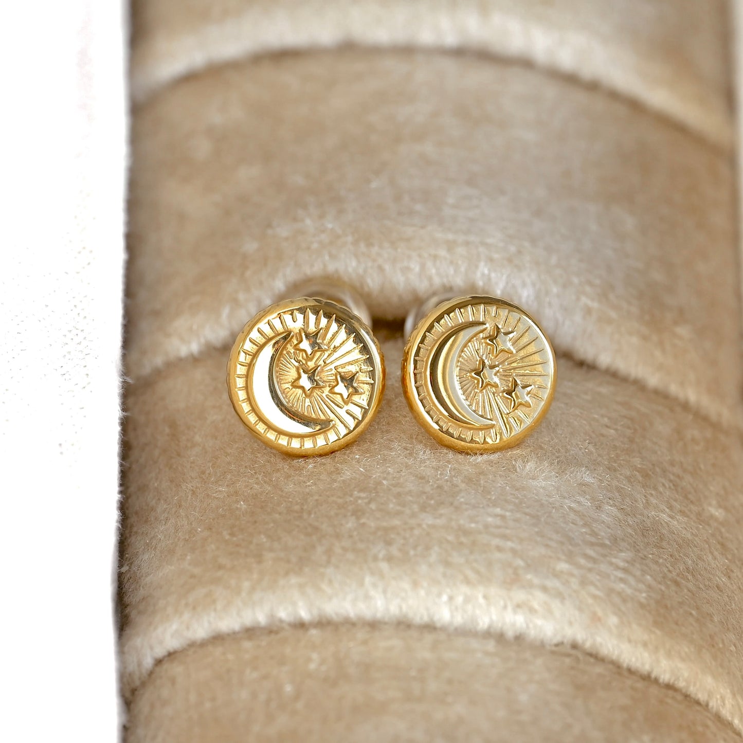 A pair of  round gold crescent moon and star stud earrings displayed on a beige velvet jewelry ring holder.