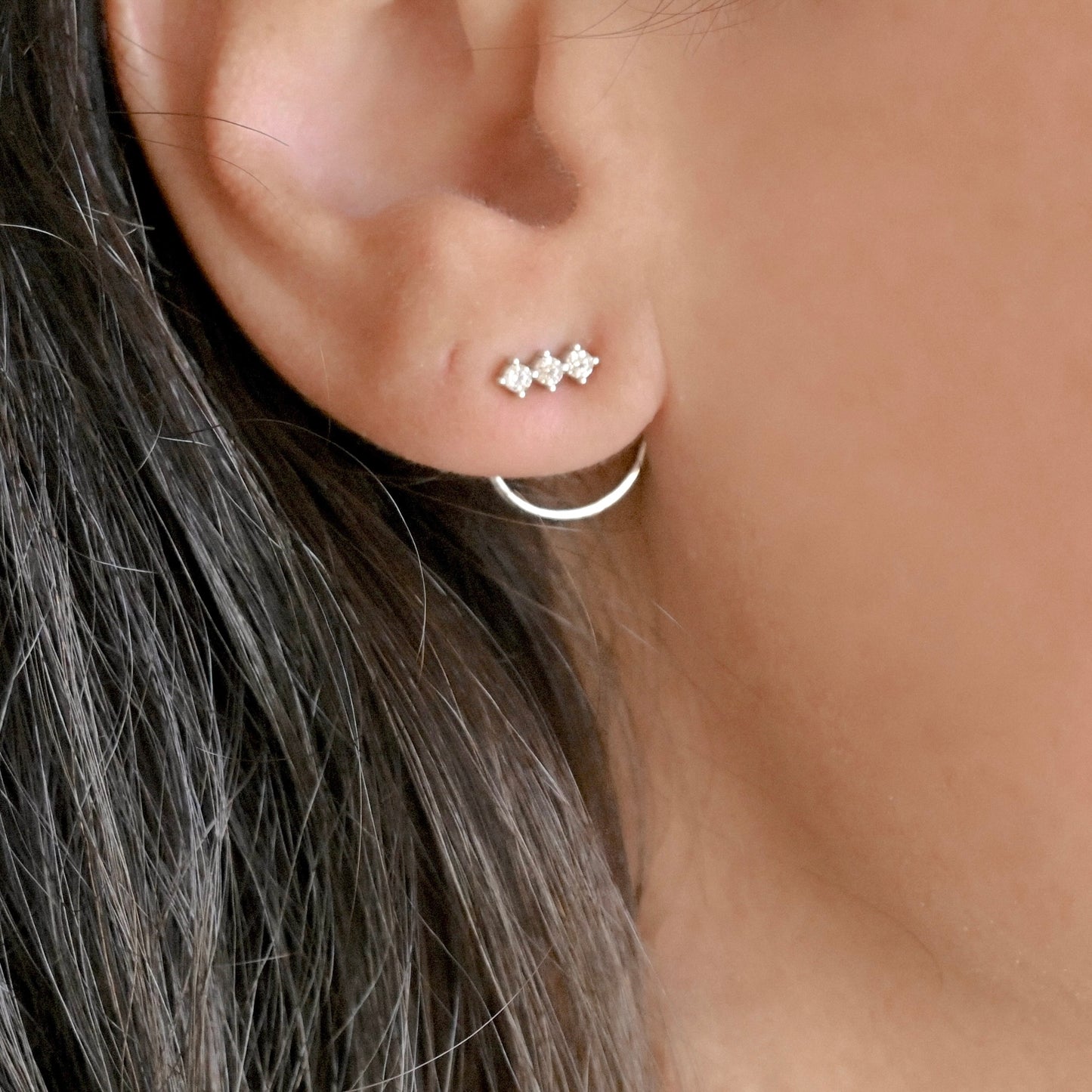 A close-up of a person's ear wearing a small silver ear jacket with three clear gemstones with a semi circle hoop at the back.