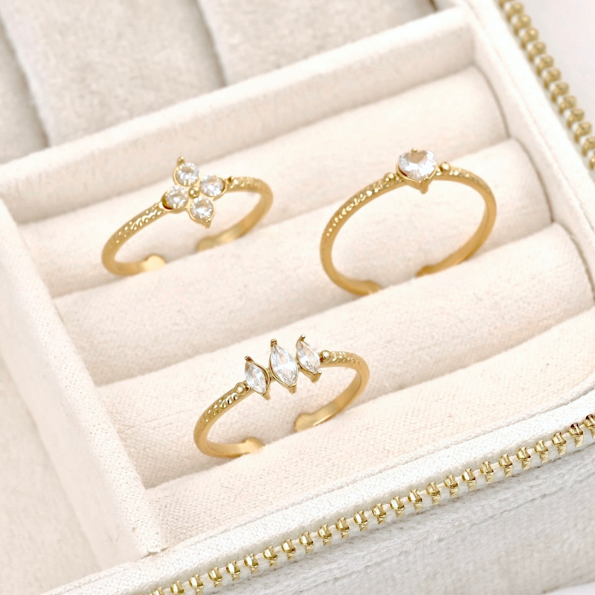 Three gold rings displayed in a cream-colored jewelry box. The rings feature clear gemstones: one has a four-stone floral cluster, one has a single marquise-cut stone, and one has a small cluster of three marquise-cut stones.