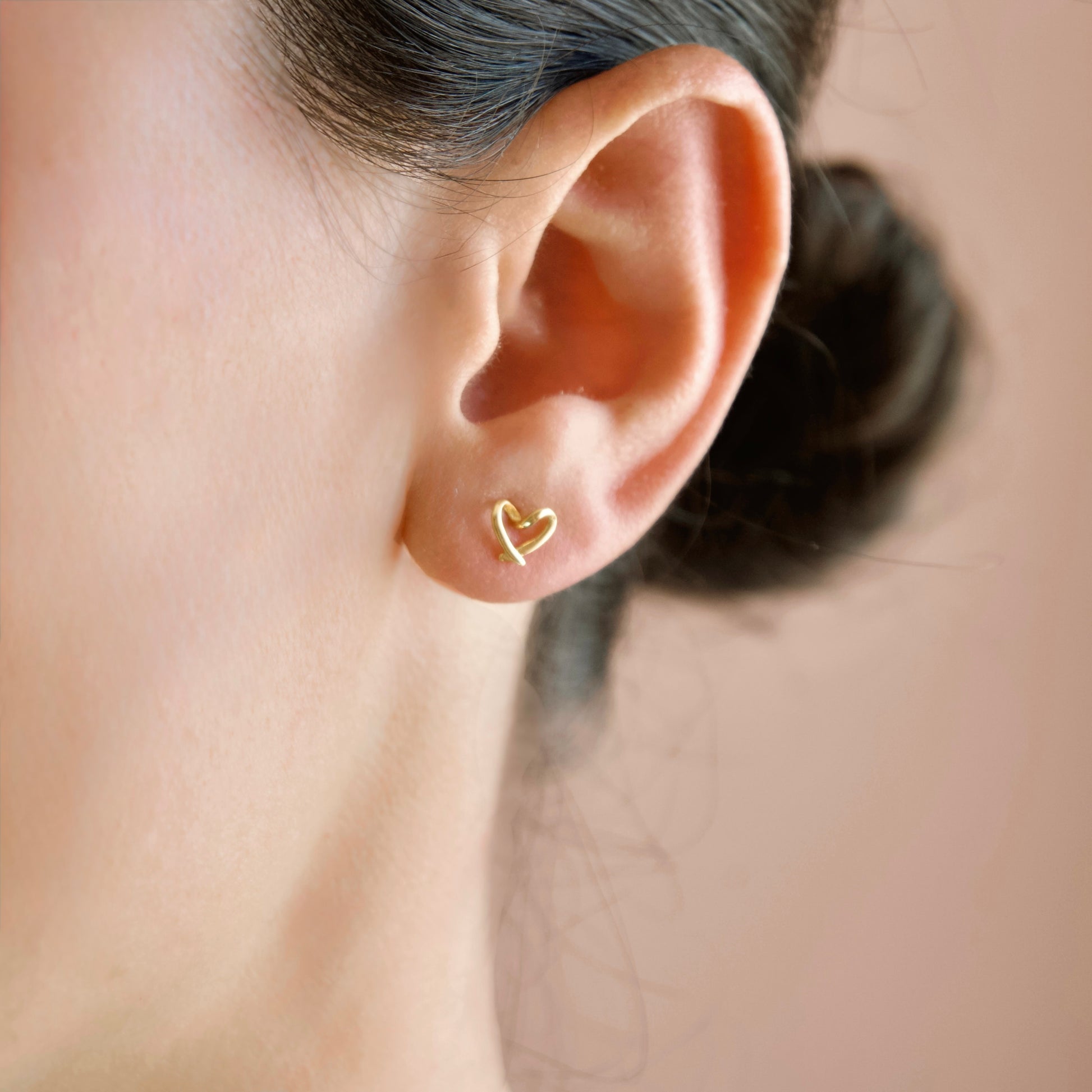 A close-up of a person's ear wearing a small open heart stud earring in polished yellow gold, set against a soft warm beige background.