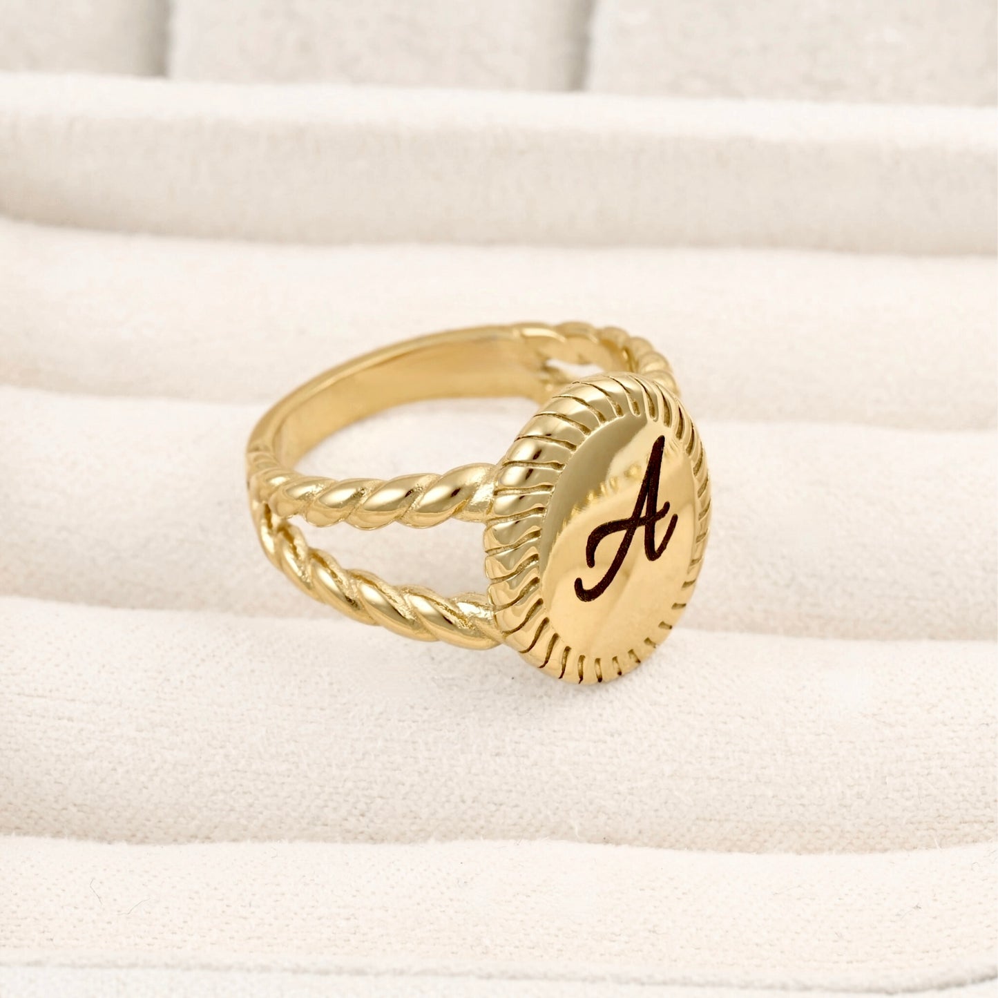 A gold signet-style ring with a twisted rope band and an oval face engraved with a dark script letter 'A', resting on a cream-colored textured jewelry display.
