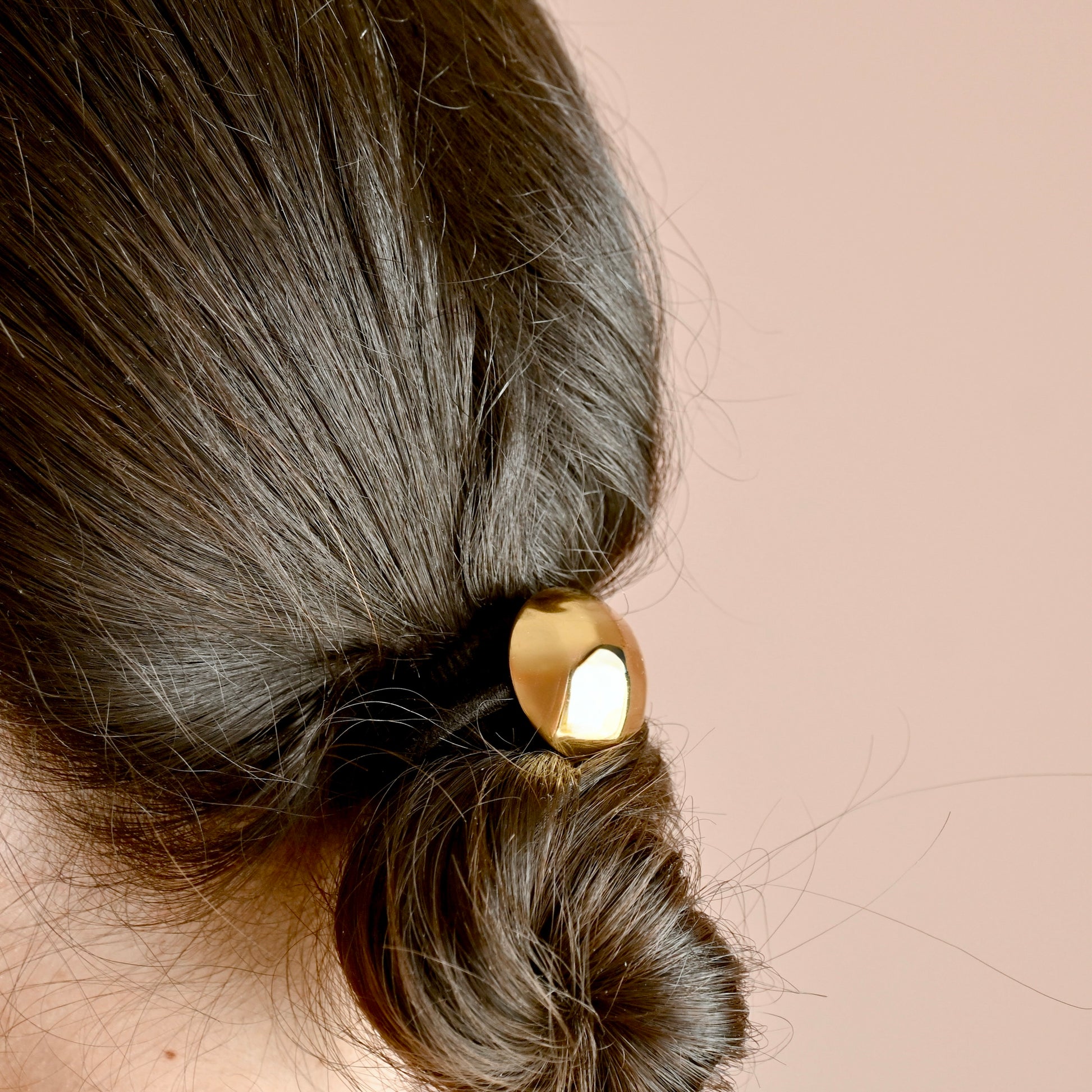 A close-up of a person's dark brown hair styled in a low bun, secured with a shiny gold dome metallic hair tie against a solid pale pink background.