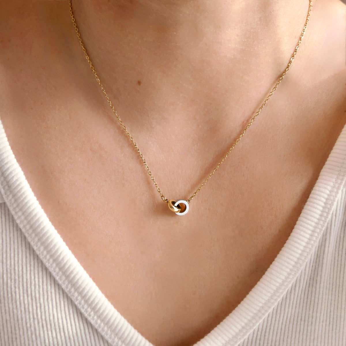 A delicate gold chain necklace with a small interlocking double-ring pendant, worn on a model's neck over a white ribbed V-neck top.